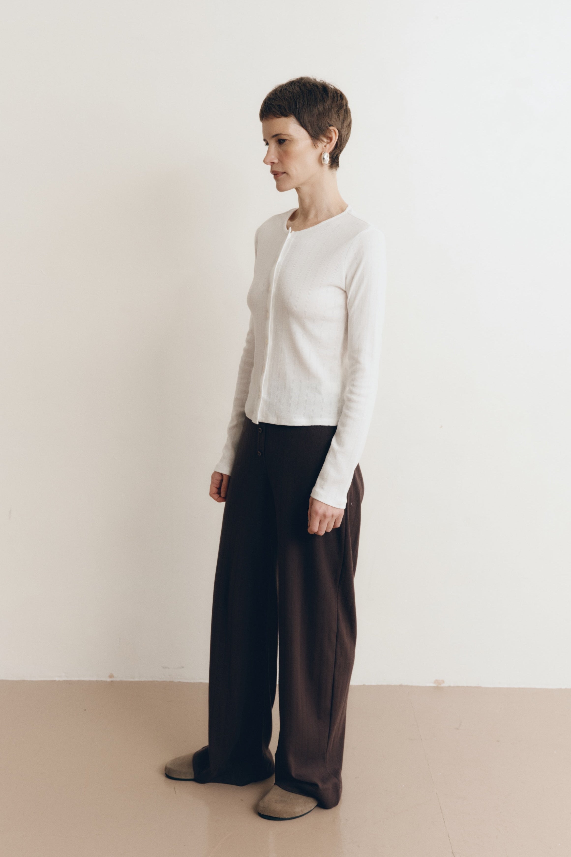 Posed female model wearing a button up sustainable 100% cotton pointelle knit cardigan in white paired with pointelle lounge pants in dark brown and brown suede shoes- VIR VITA
#color_coconut