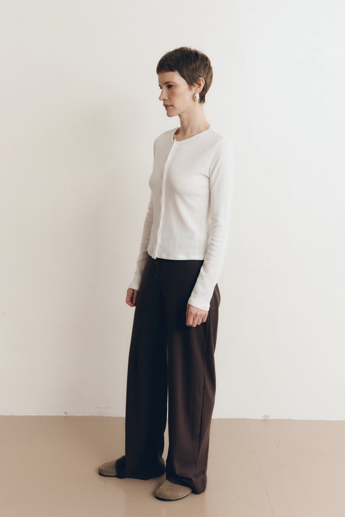 Posed female model wearing a button up sustainable 100% cotton pointelle knit cardigan in white paired with pointelle lounge pants in dark brown and brown suede shoes- VIR VITA
#color_coconut