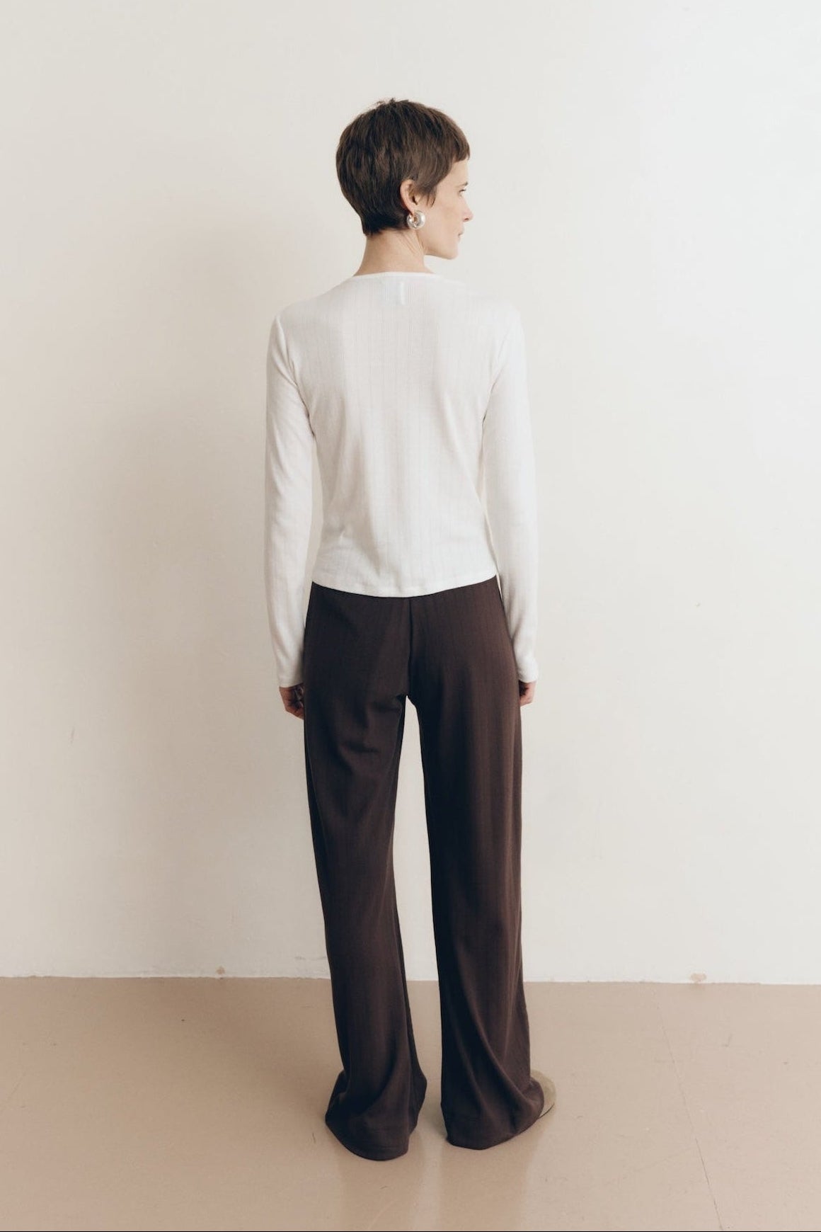 Posed female model wearing a button up sustainable 100% cotton pointelle knit cardigan in white paired with pointelle lounge pants in dark brown and brown suede shoes- VIR VITA
#color_coconut