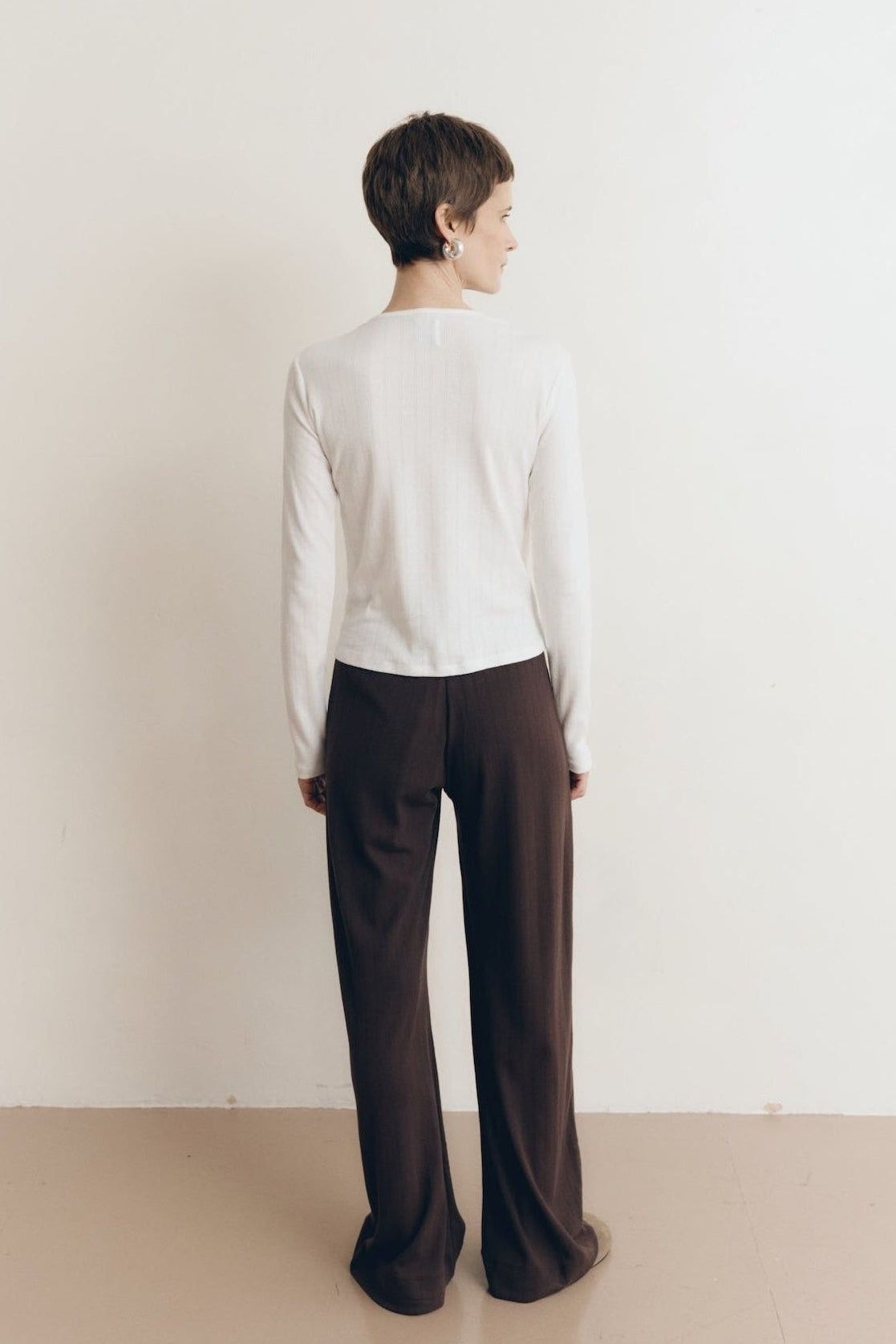 Posed female model wearing a button up sustainable 100% cotton pointelle knit cardigan in white paired with pointelle lounge pants in dark brown and brown suede shoes- VIR VITA
#color_coconut