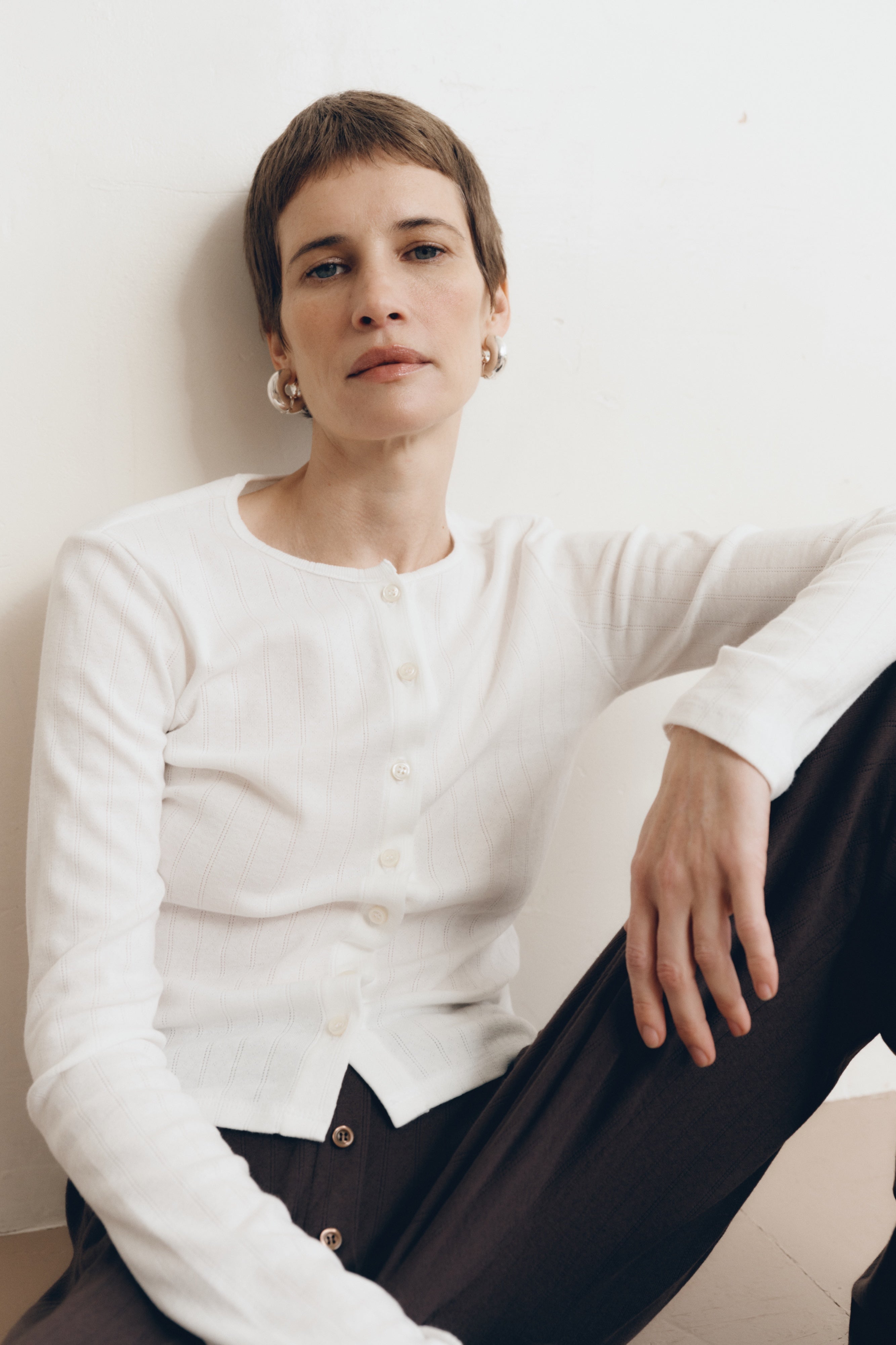 Posed female model wearing a button up sustainable 100% cotton pointelle knit cardigan in white paired with pointelle lounge pants in dark brown- VIR VITA
#color_coconut