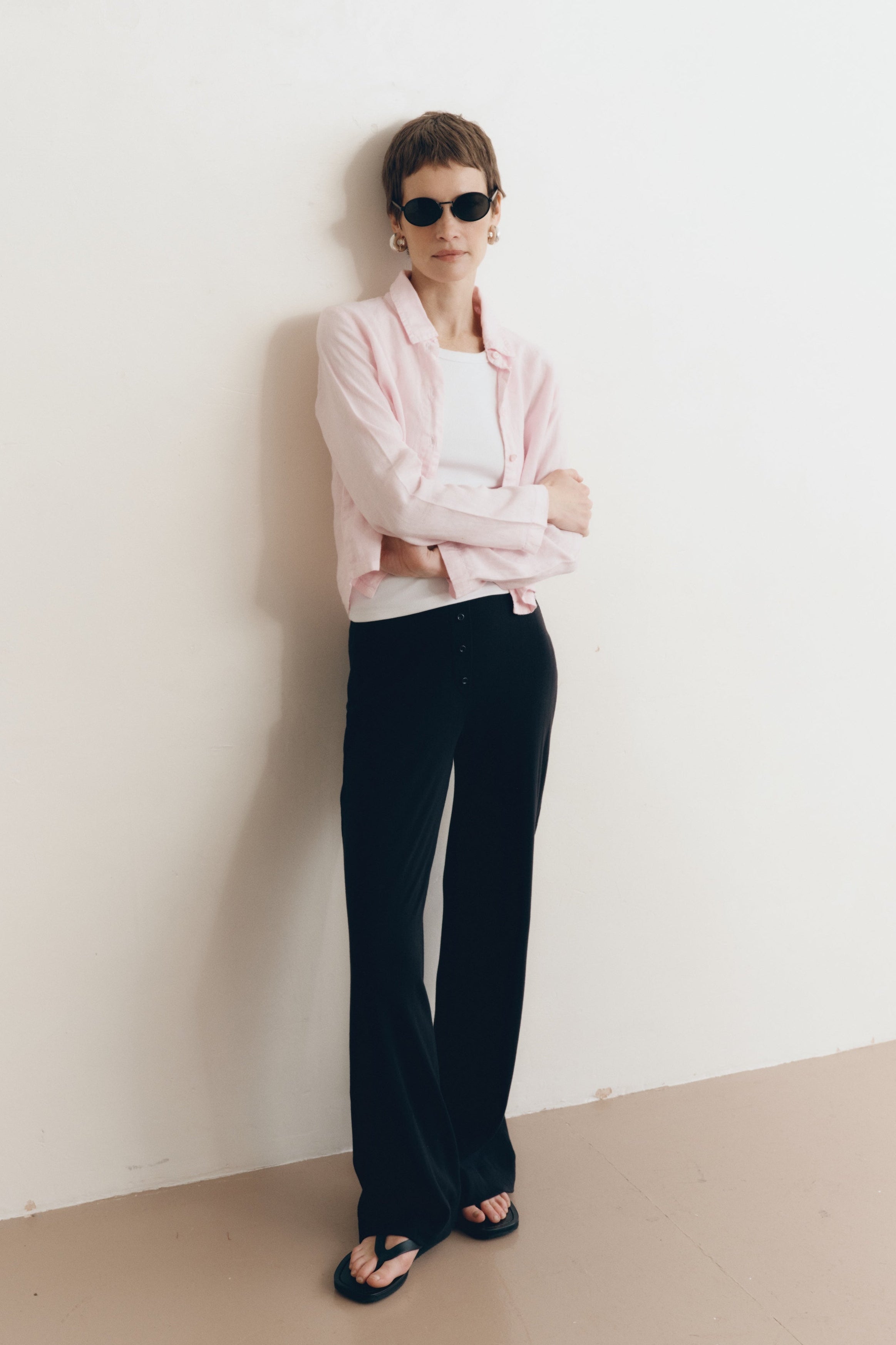 Posed female model wearing a sustainable 100% linen classic button up collared shirt in light pink paired with a high neck white tank top, black pointelle lounge pants, black sunglassess, and black sandals- VIR VITA
#color_blossom
