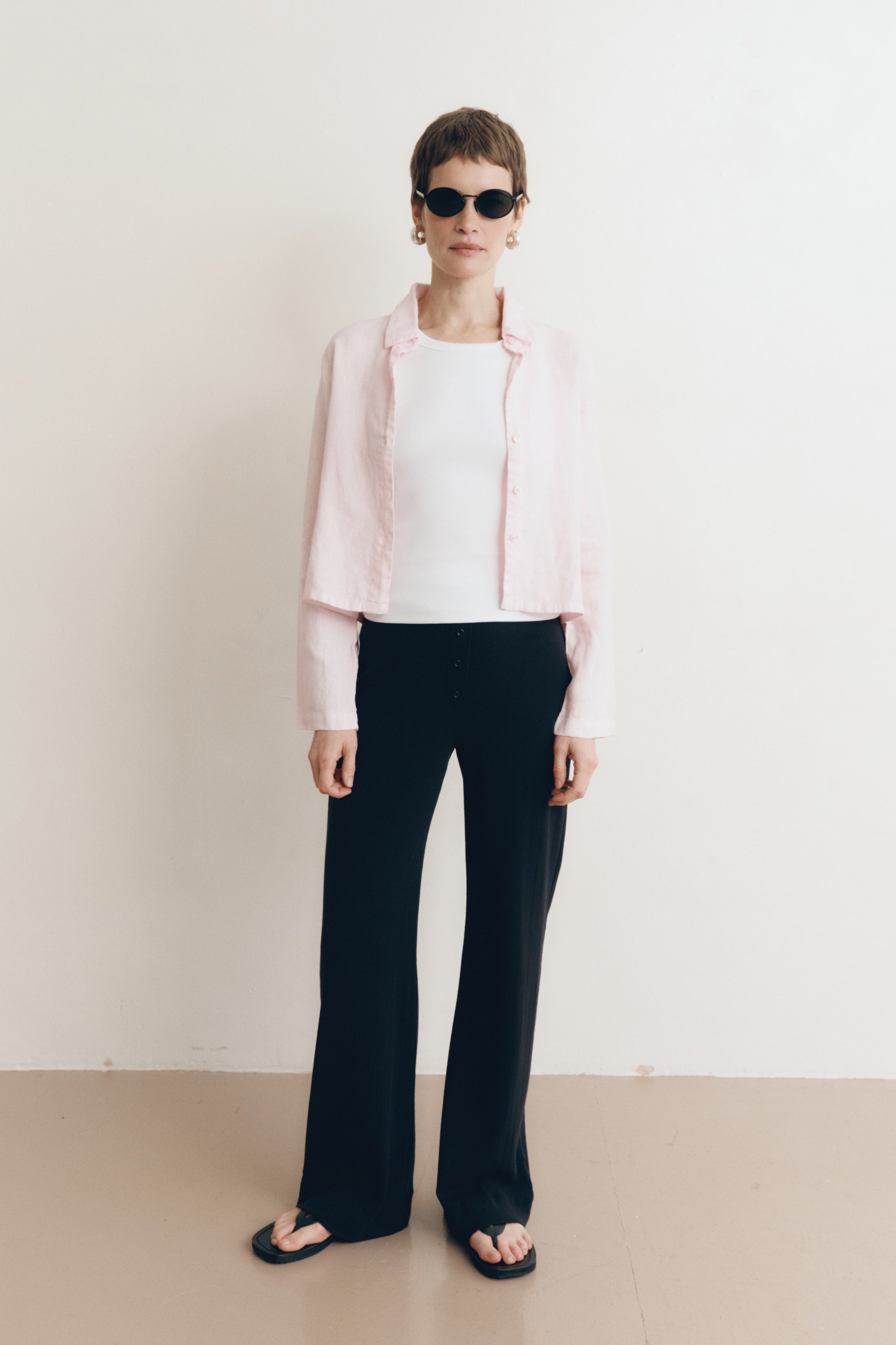 Posed female model wearing a sustainable 100% linen classic button up collared shirt in light pink paired with a high neck white tank top, black pointelle lounge pants, black sunglassess, and black sandals- VIR VITA
#color_blossom