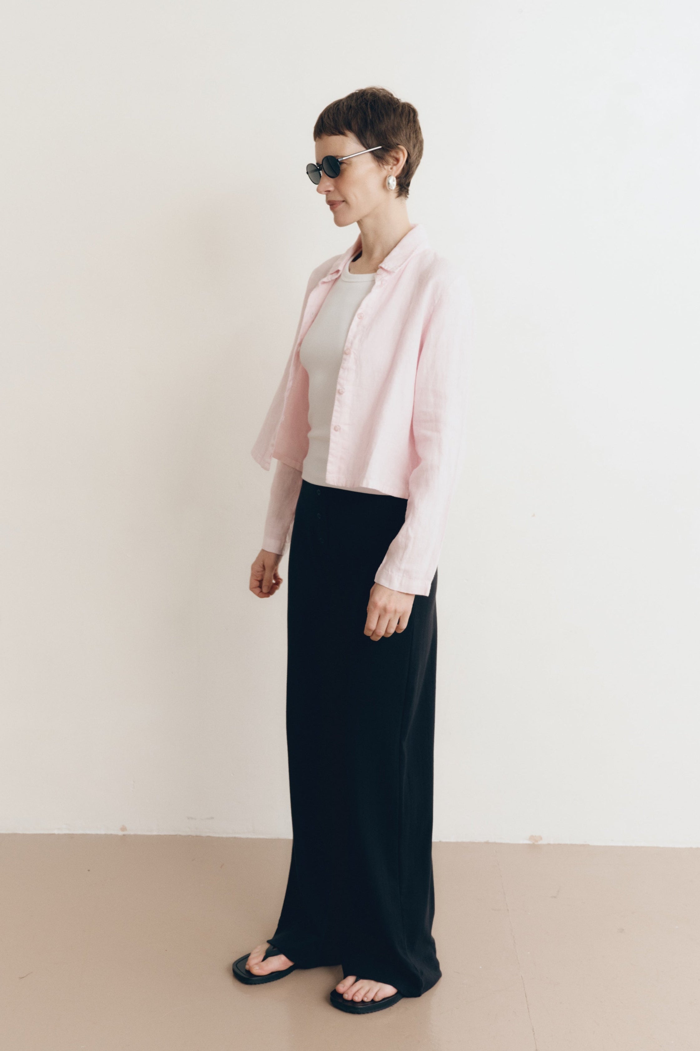 Posed female model wearing a sustainable 100% linen classic button up collared shirt in light pink paired with a high neck white tank top, black pointelle lounge pants, black sunglassess, and black sandals- VIR VITA
#color_blossom