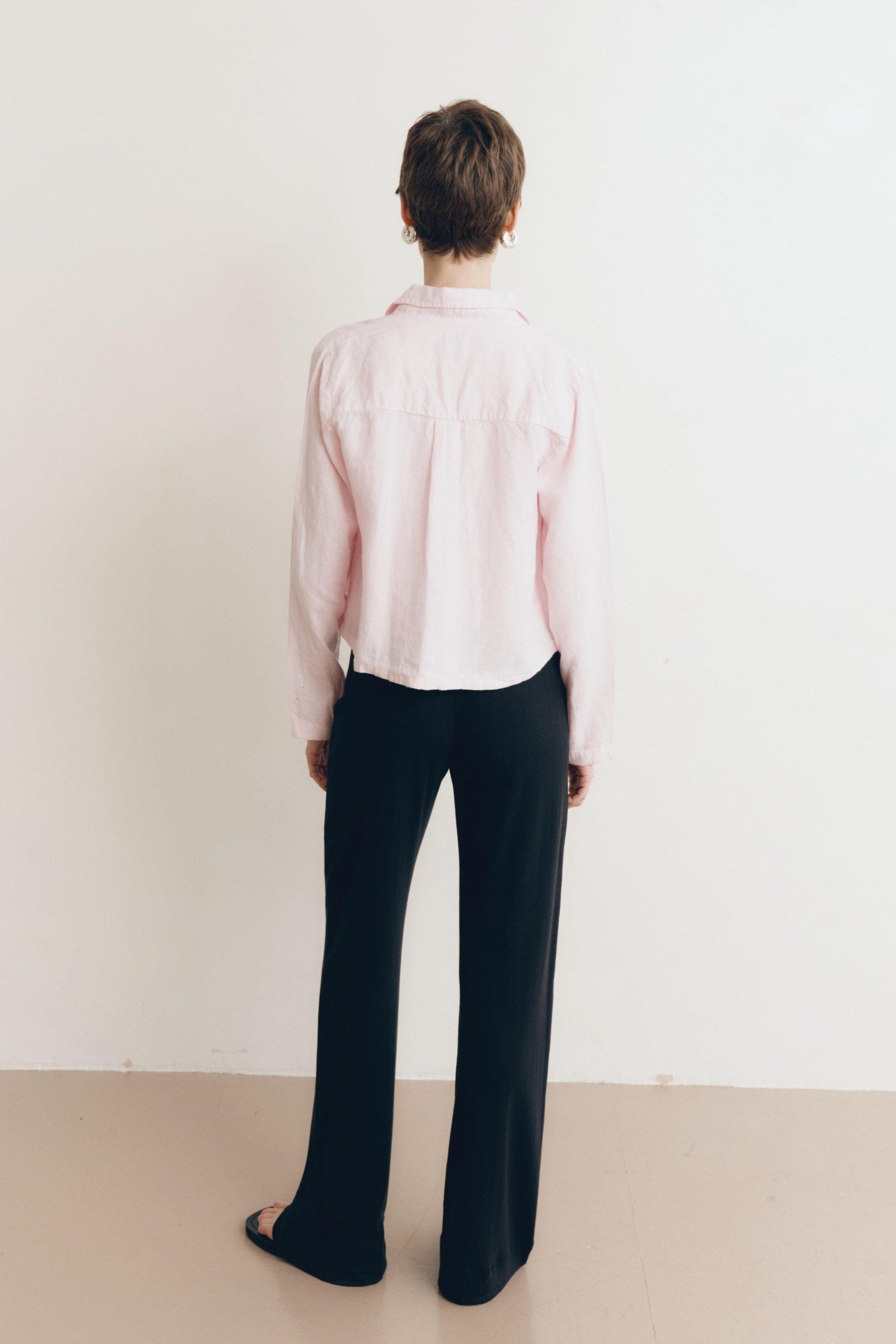 Posed female model wearing a sustainable 100% linen classic button up collared shirt in light pink paired with a high neck white tank top, black pointelle lounge pants, and black sandals- VIR VITA
#color_blossom