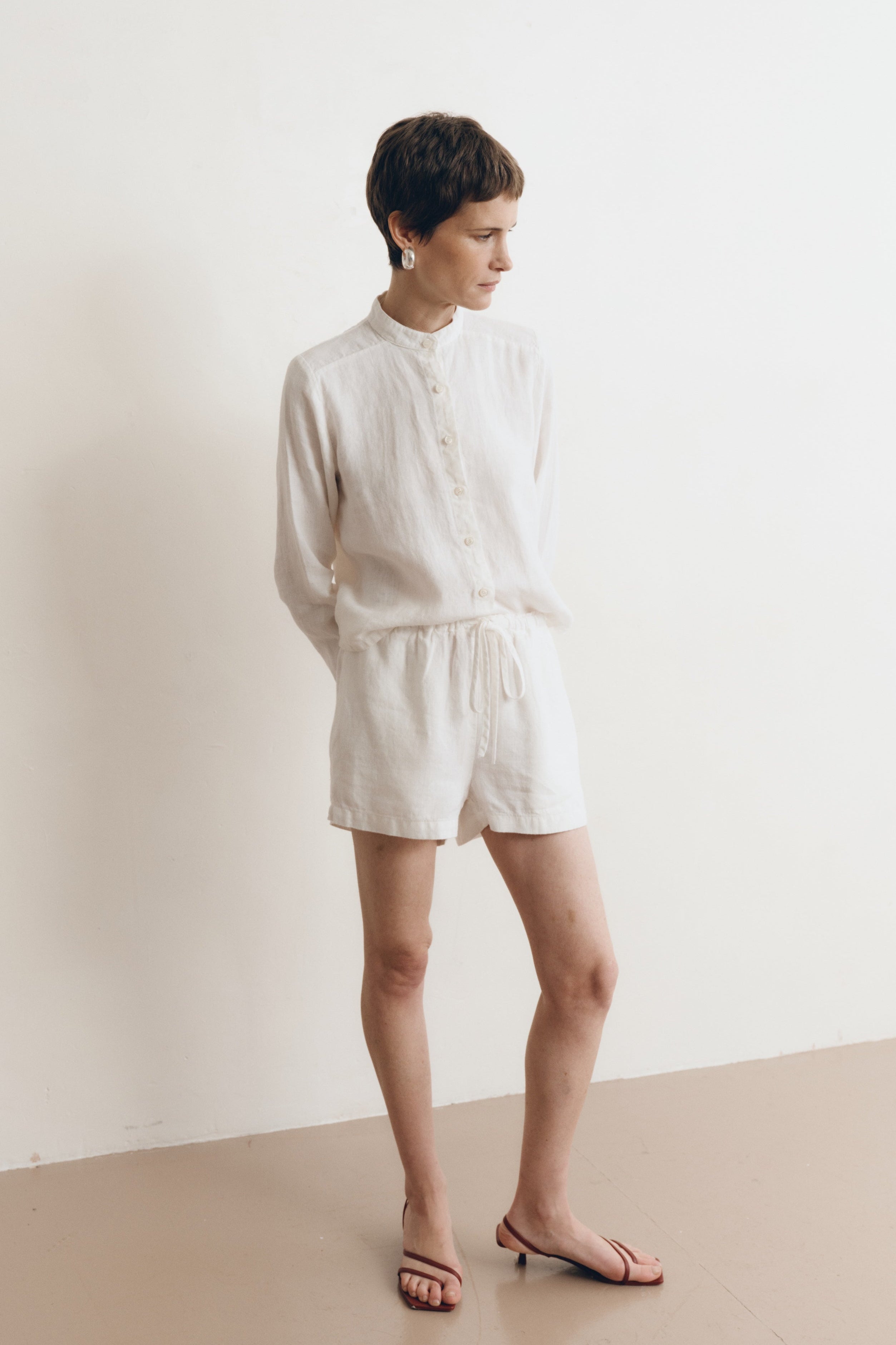 Posed female model wearing a white button up 100% linen shirt with a band collar and cropped silhouette paired with matching white linen drawstring shorts, and brown heels - VIR VITA
#color_white