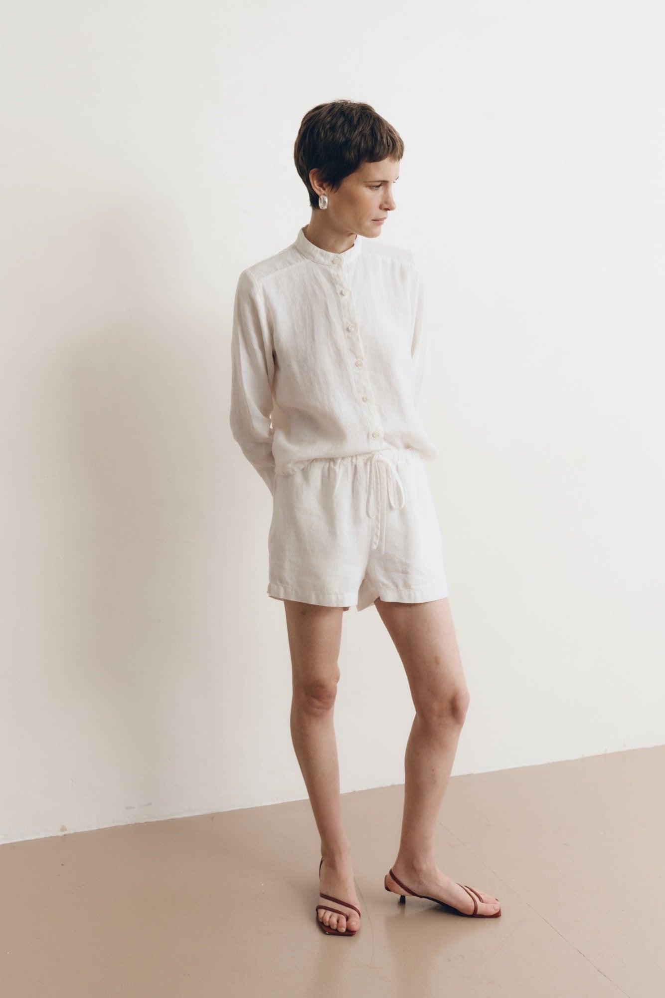 Posed female model wearing sustainable 100% linen drawstring shorts in white paired with a linen button up oversized shirt in white, and red sandals - VIR VITA
#color_white