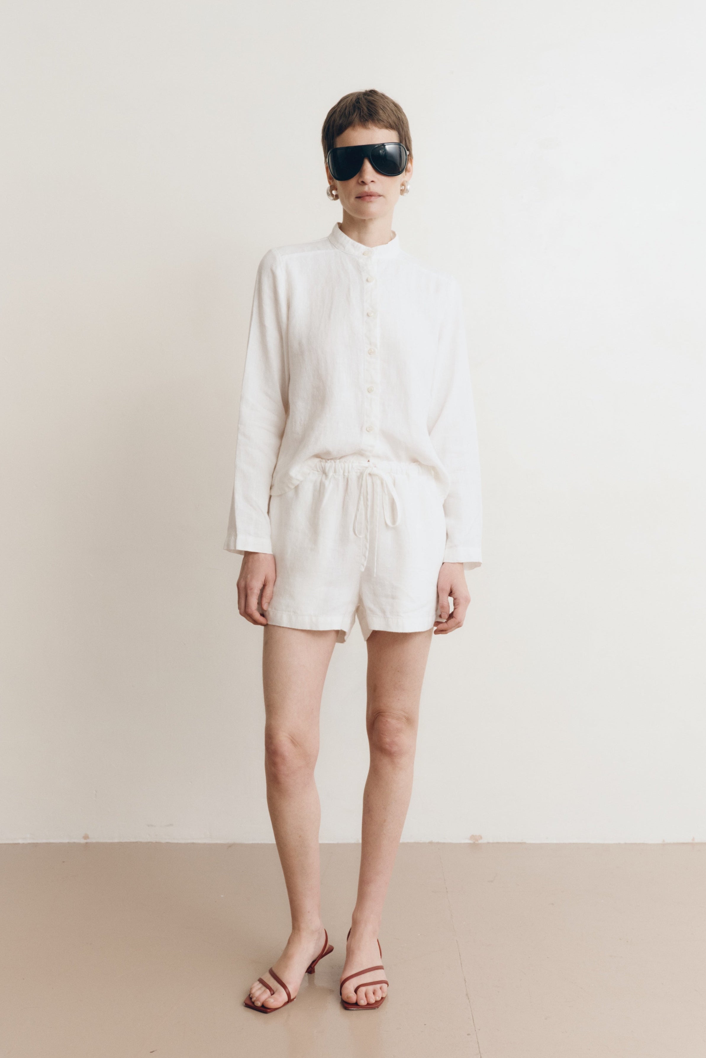 Posed female model wearing a white button up 100% linen shirt with a band collar and cropped silhouette paired with matching white linen drawstring shorts, brown heels, and black sunglasses - VIR VITA
#color_white