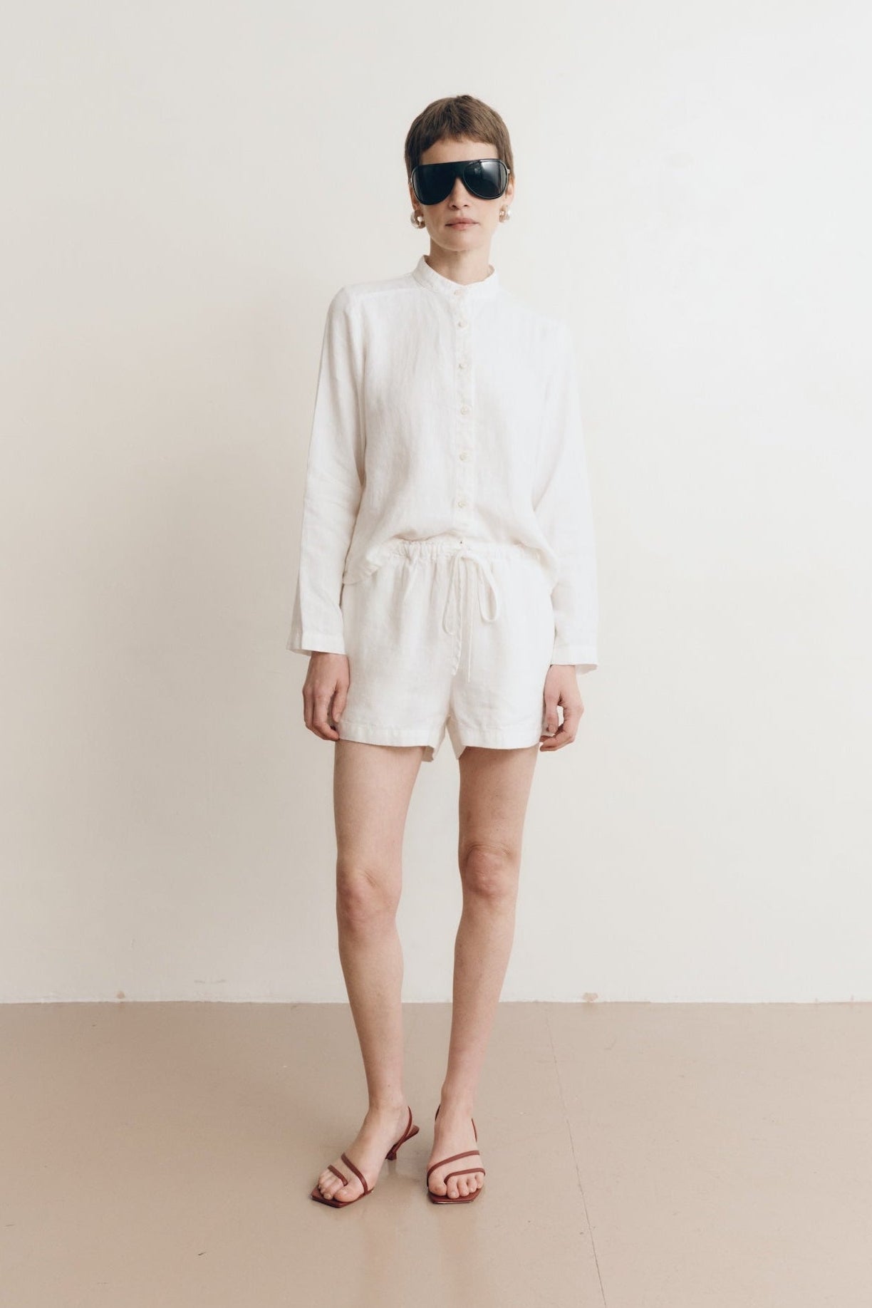 Posed female model wearing sustainable 100% linen drawstring shorts in white paired with a linen button up oversized shirt in white, and red sandals - VIR VITA
#color_white