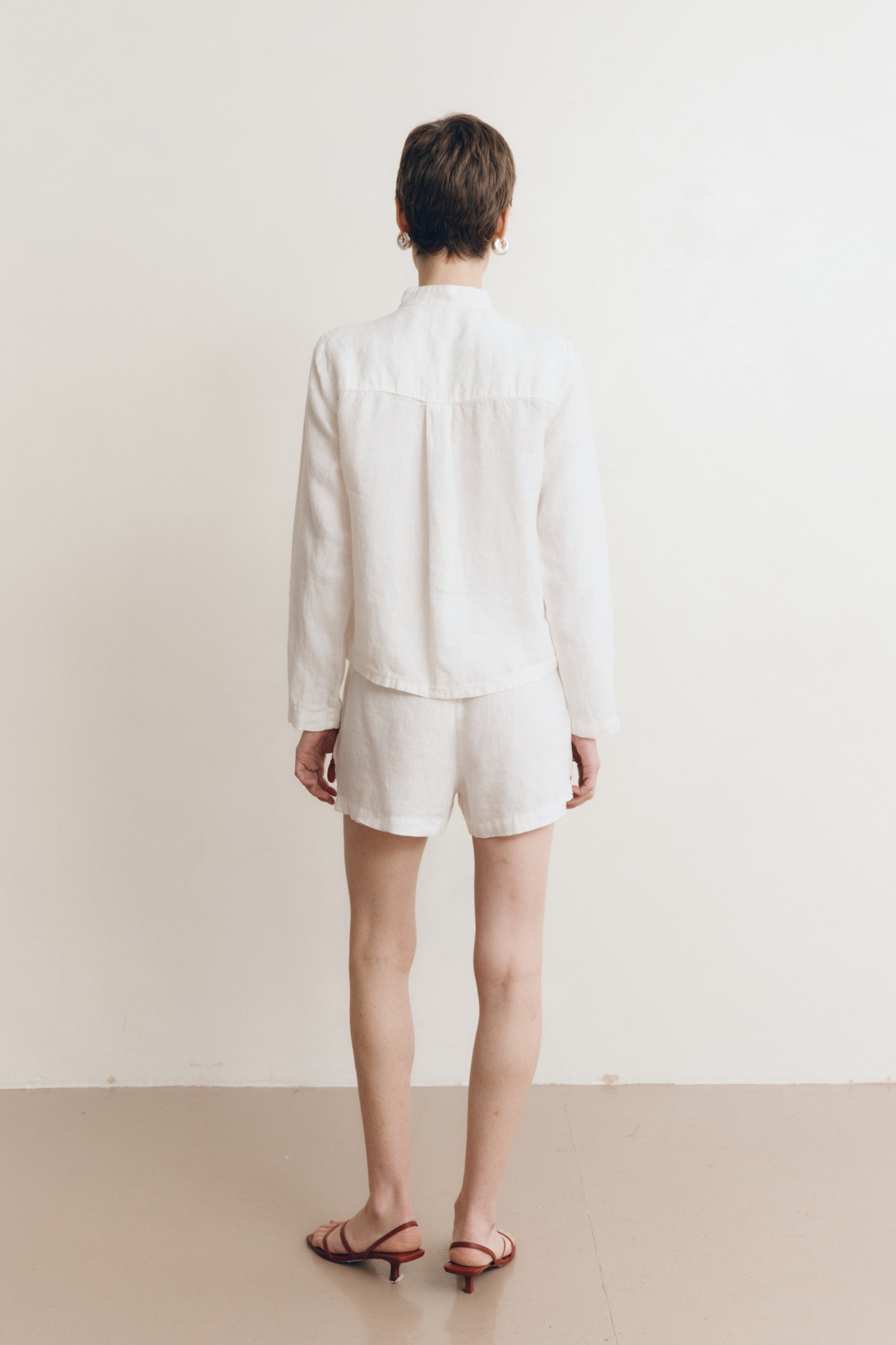 Posed female model wearing a white button up 100% linen shirt with a band collar and cropped silhouette paired with matching white linen drawstring shorts, and brown heels- VIR VITA
#color_white
