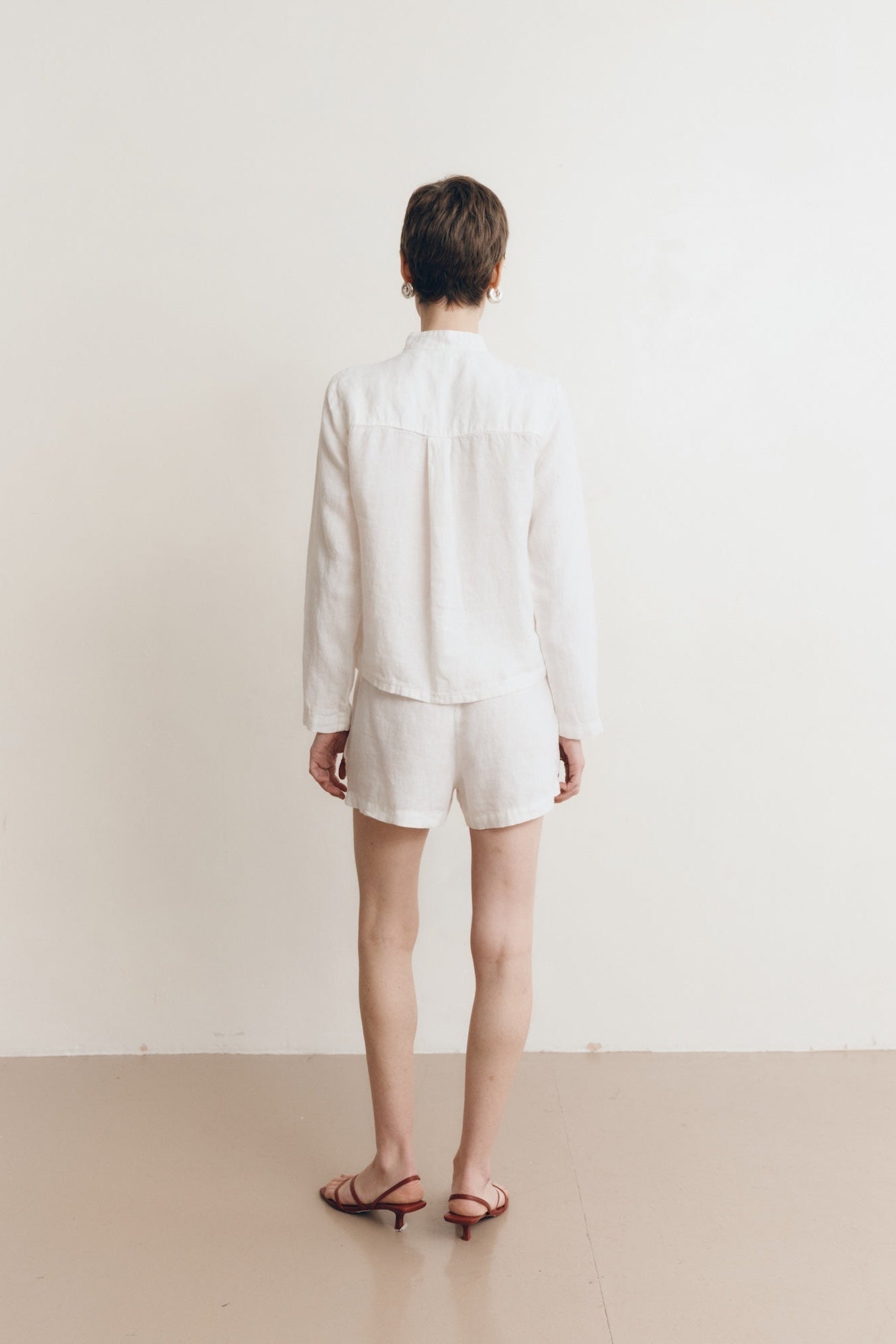 Posed female model wearing sustainable 100% linen drawstring shorts in white paired with a linen button up oversized shirt in white, and red sandals - VIR VITA
#color_white