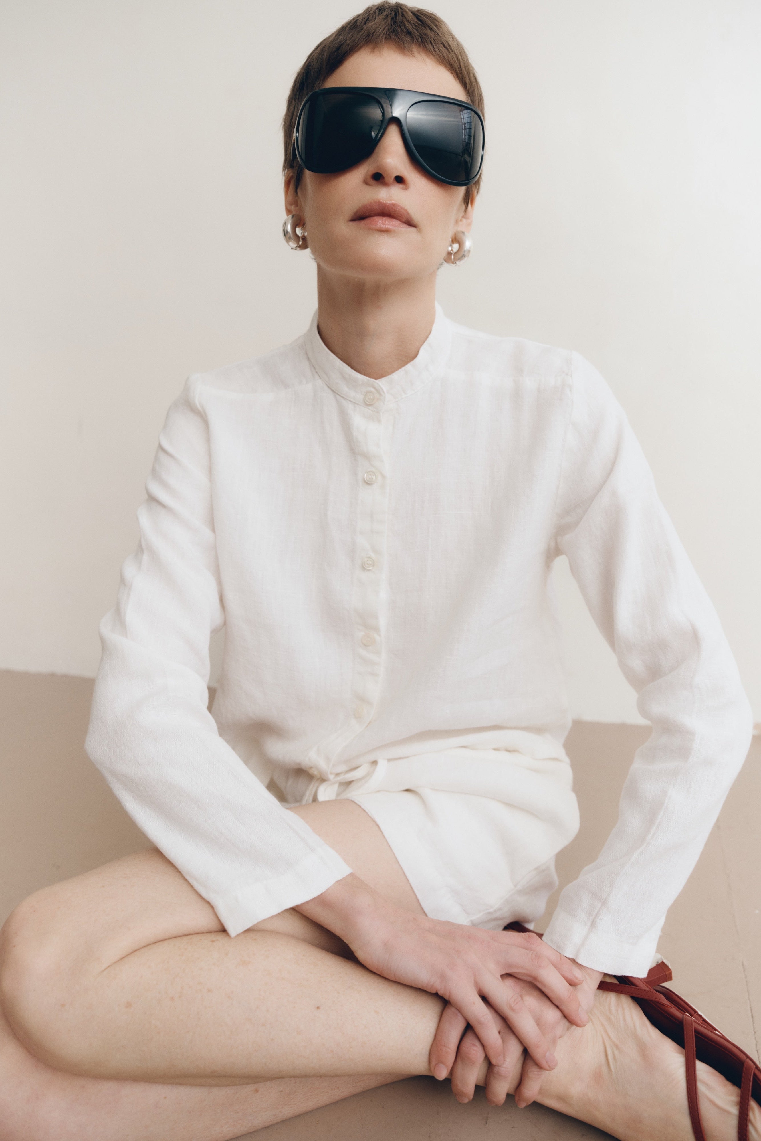 Posed female model wearing a white button up 100% linen shirt with a band collar and cropped silhouette paired with matching white linen drawstring shorts, brown heels, and black sunglasses - VIR VITA
#color_white