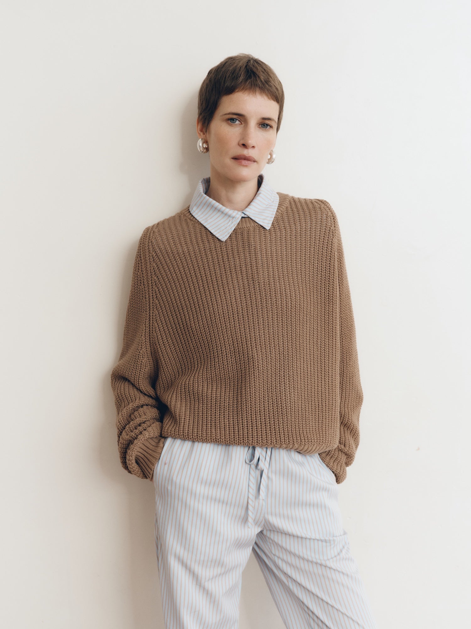 Posed female model wearing a 100% cotton knit sweater with a flattering and subtly cropped silhouette in light brown paired with a striped collared shirt in light blue - VIR VITA
#color_bark
