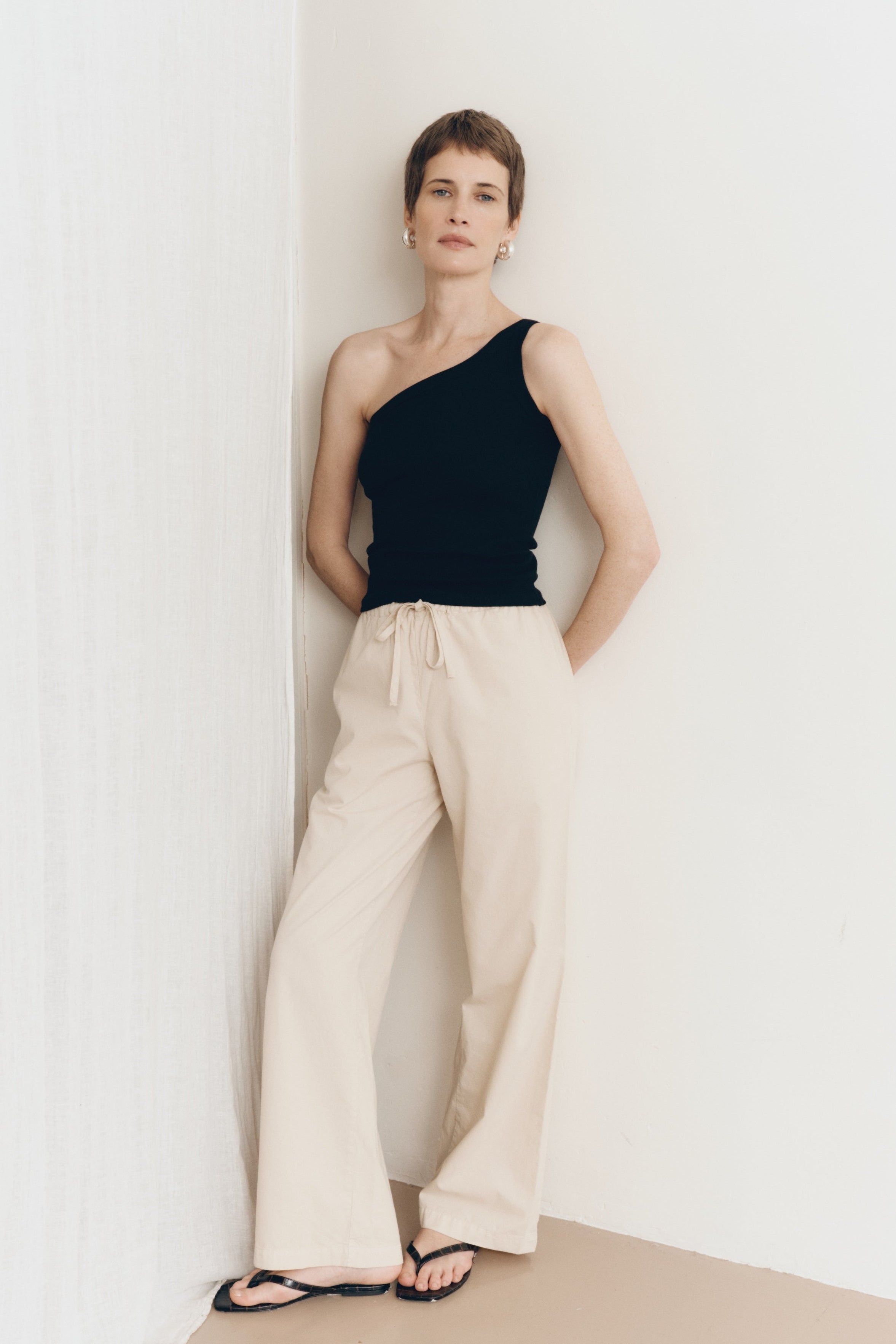 Posed female model wearing 100% cotton poplin drawstring relaxed pants in beige paired with a one shoulder tank top in black and black sandals - VIR VITA
#color_dune