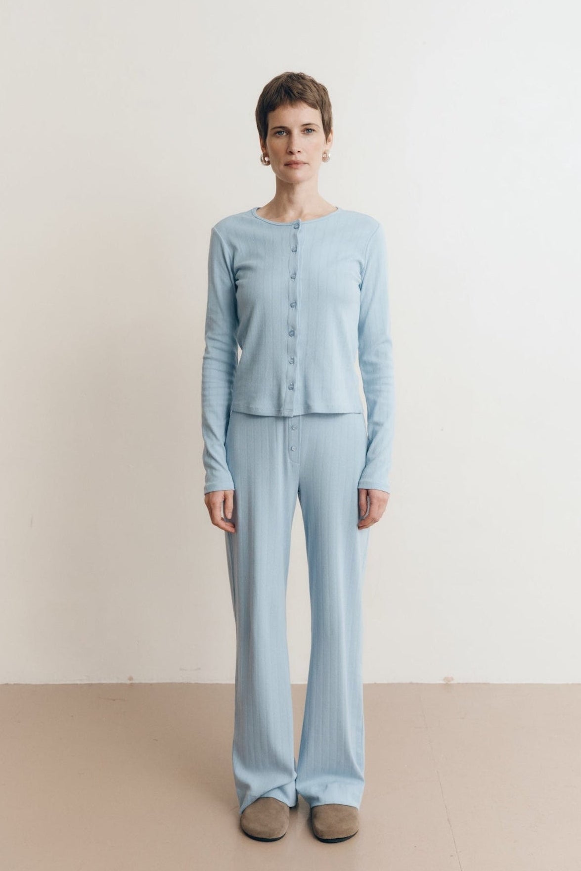 Posed female model wearing a button up sustainable 100% cotton pointelle knit cardigan in light blue paired with a matching pointelle scoop neck tee and pointelle lounge pants with brown suede shoes- VIR VITA
#color_vintage blue