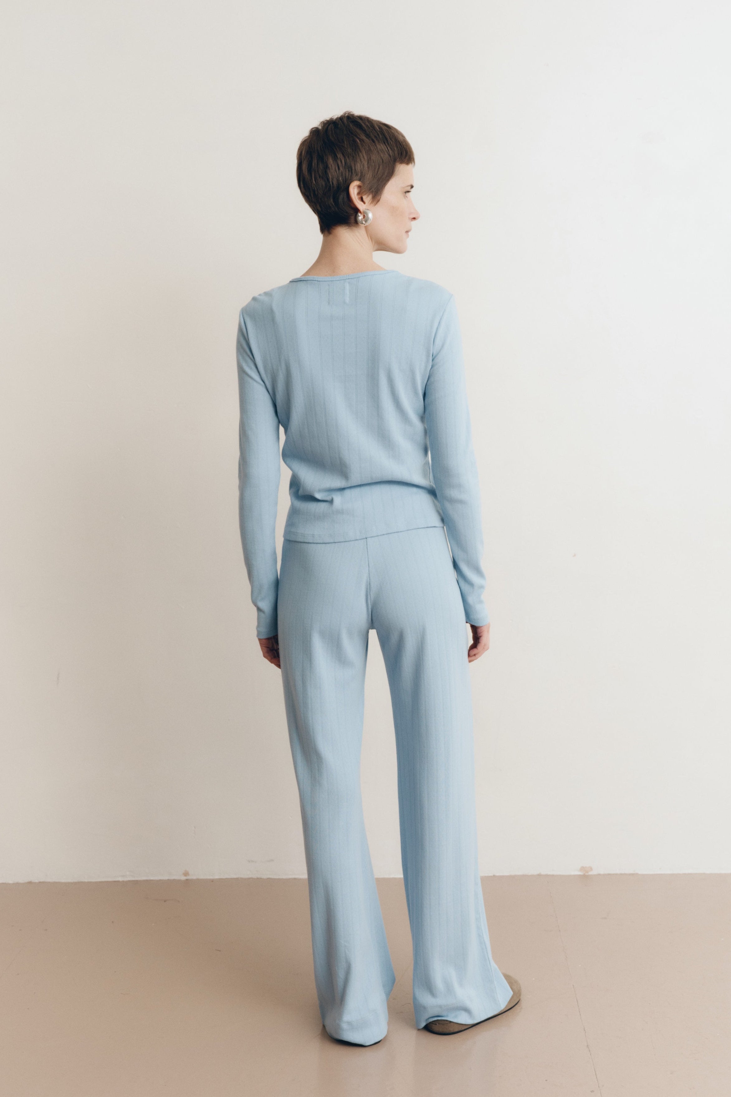 Posed female model wearing a button up sustainable 100% cotton pointelle knit cardigan in light blue paired with matching pointelle lounge pants with brown suede shoes- VIR VITA
#color_vintage blue