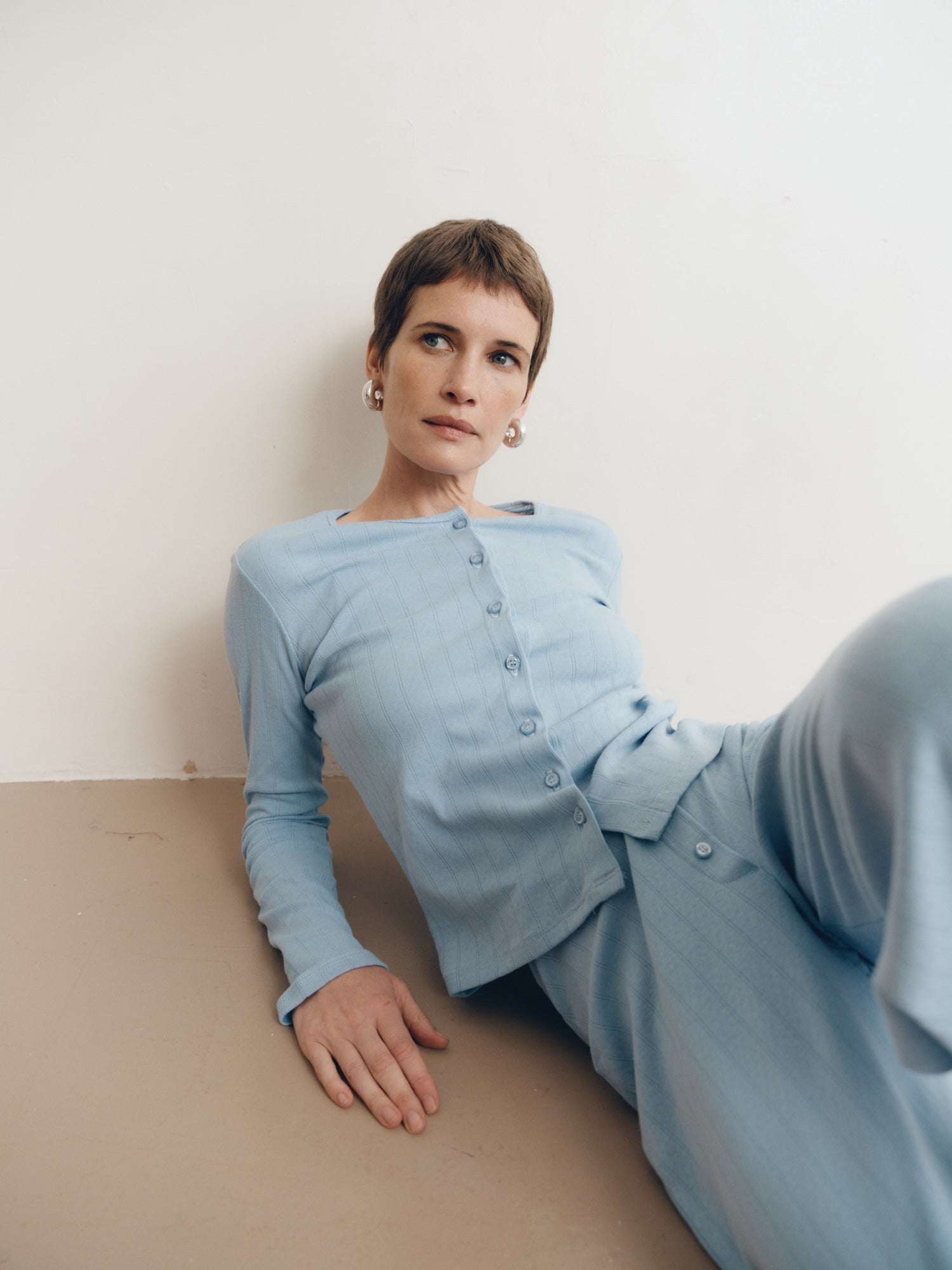 Posed female model wearing a button up sustainable 100% cotton pointelle knit cardigan in light blue paired with matching pointelle knit lounge pants and brown suede shoes - VIR VITA
#color_vintage blue