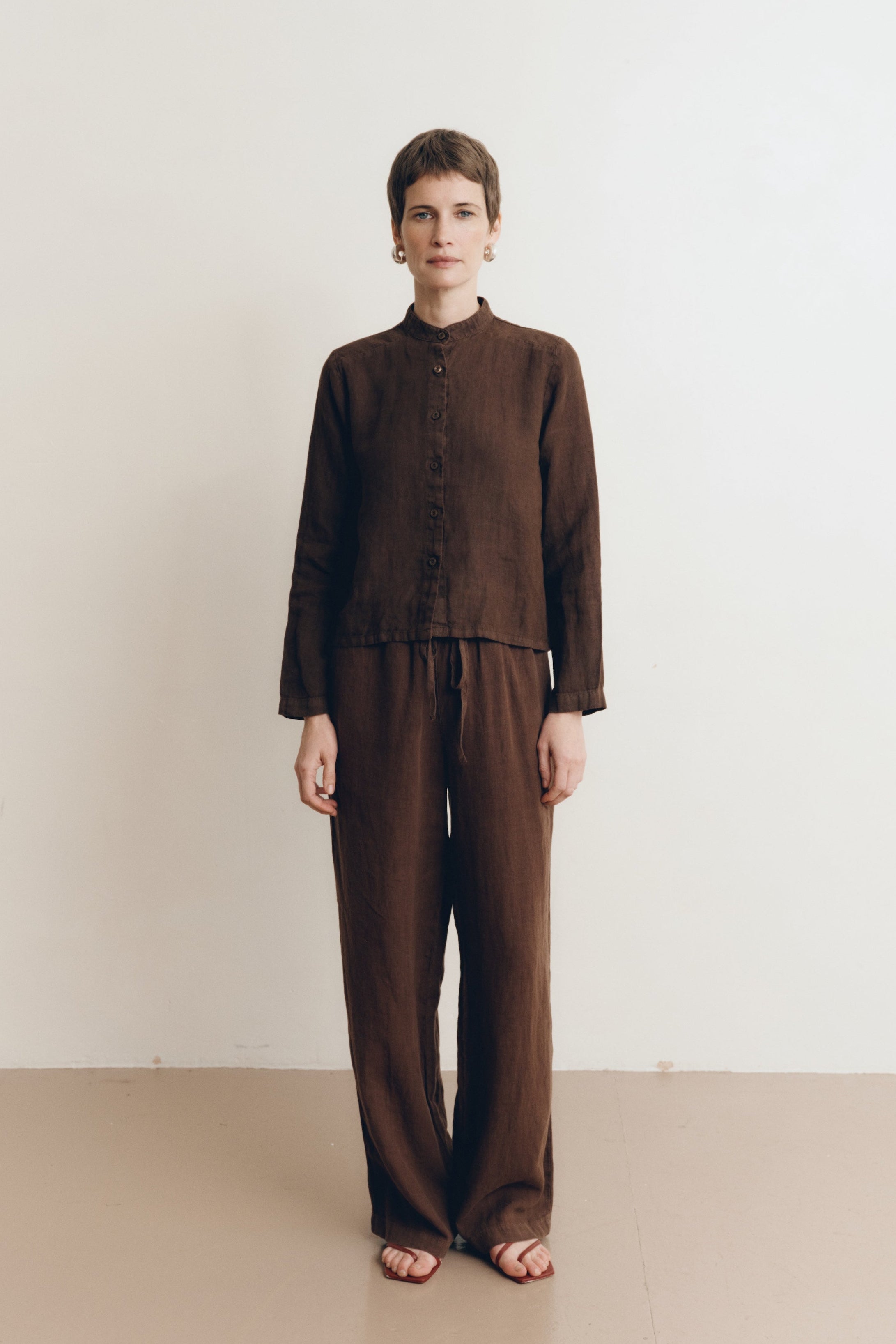 Posed female model wearing a dark brown button up 100% linen shirt with a band collar and cropped silhouette paired with matching dark brown linen drawstring pants, and brown heels - VIR VITA
#color_chestnut