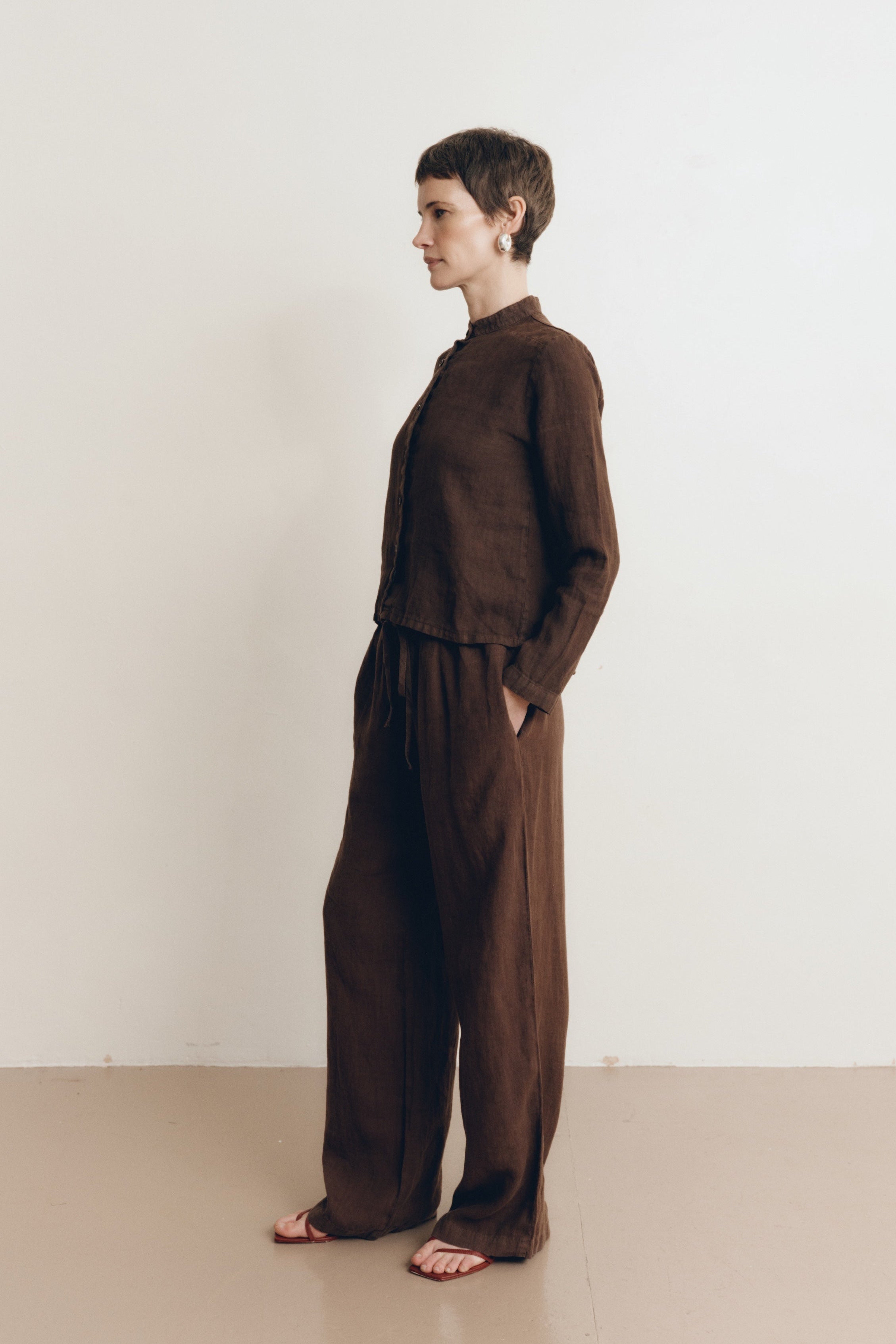 Posed female model wearing a dark brown button up 100% linen shirt with a band collar and cropped silhouette paired with matching dark brown linen drawstring pants, and brown heels- VIR VITA
#color_chestnut