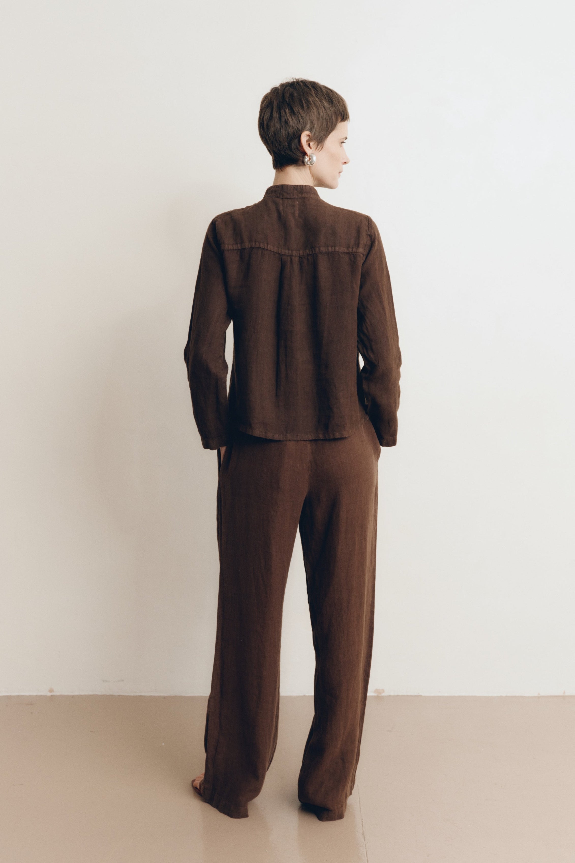 Posed female model wearing a dark brown button up 100% linen shirt with a band collar and cropped silhouette paired with matching dark brown linen drawstring pants, and brown heels - VIR VITA
#color_chestnut