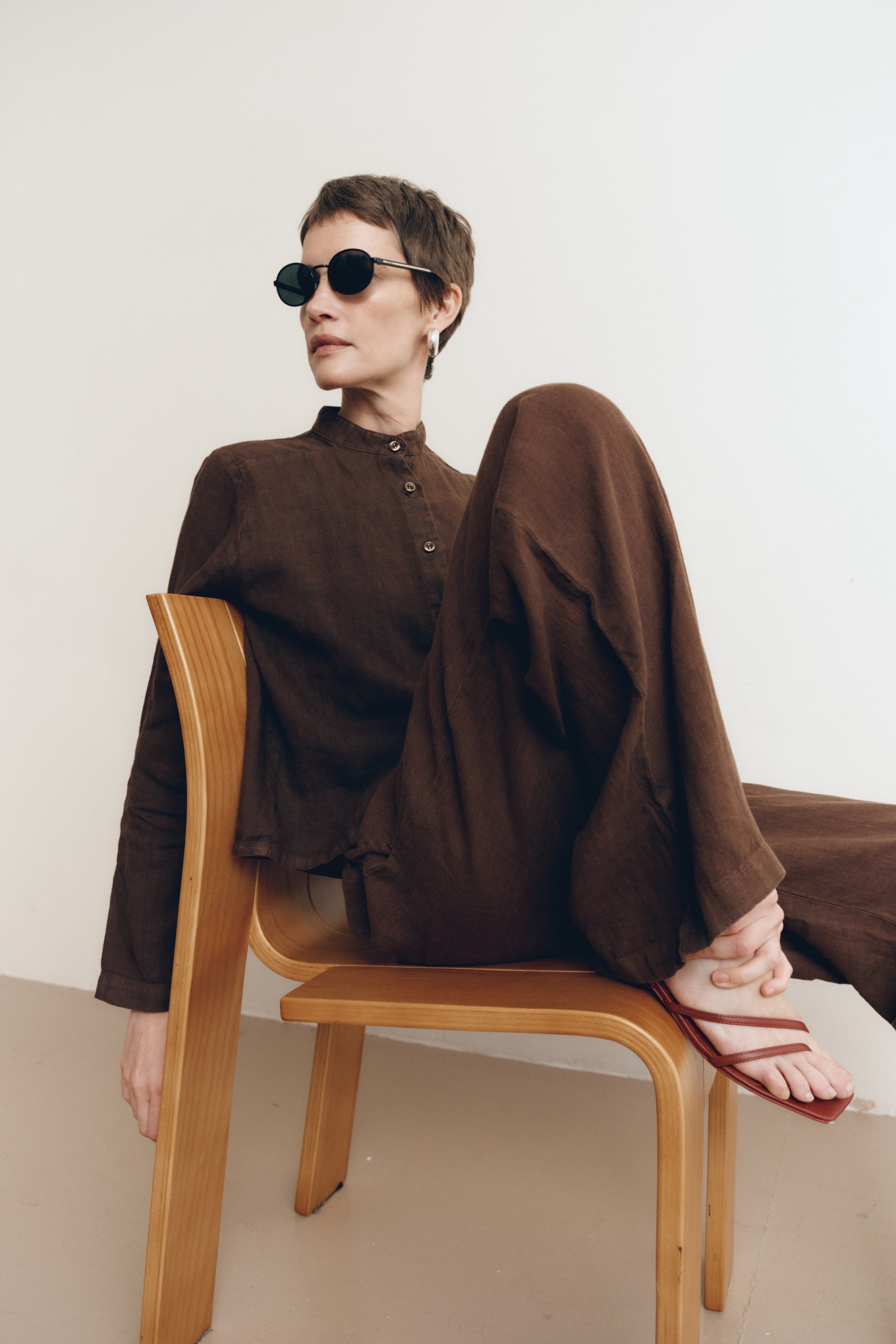 Posed female model sitting on a chair wearing a dark brown button up 100% linen shirt with a band collar and cropped silhouette paired with matching dark brown linen drawstring pants, brown heels, and black sunglasses - VIR VITA
#color_chestnut