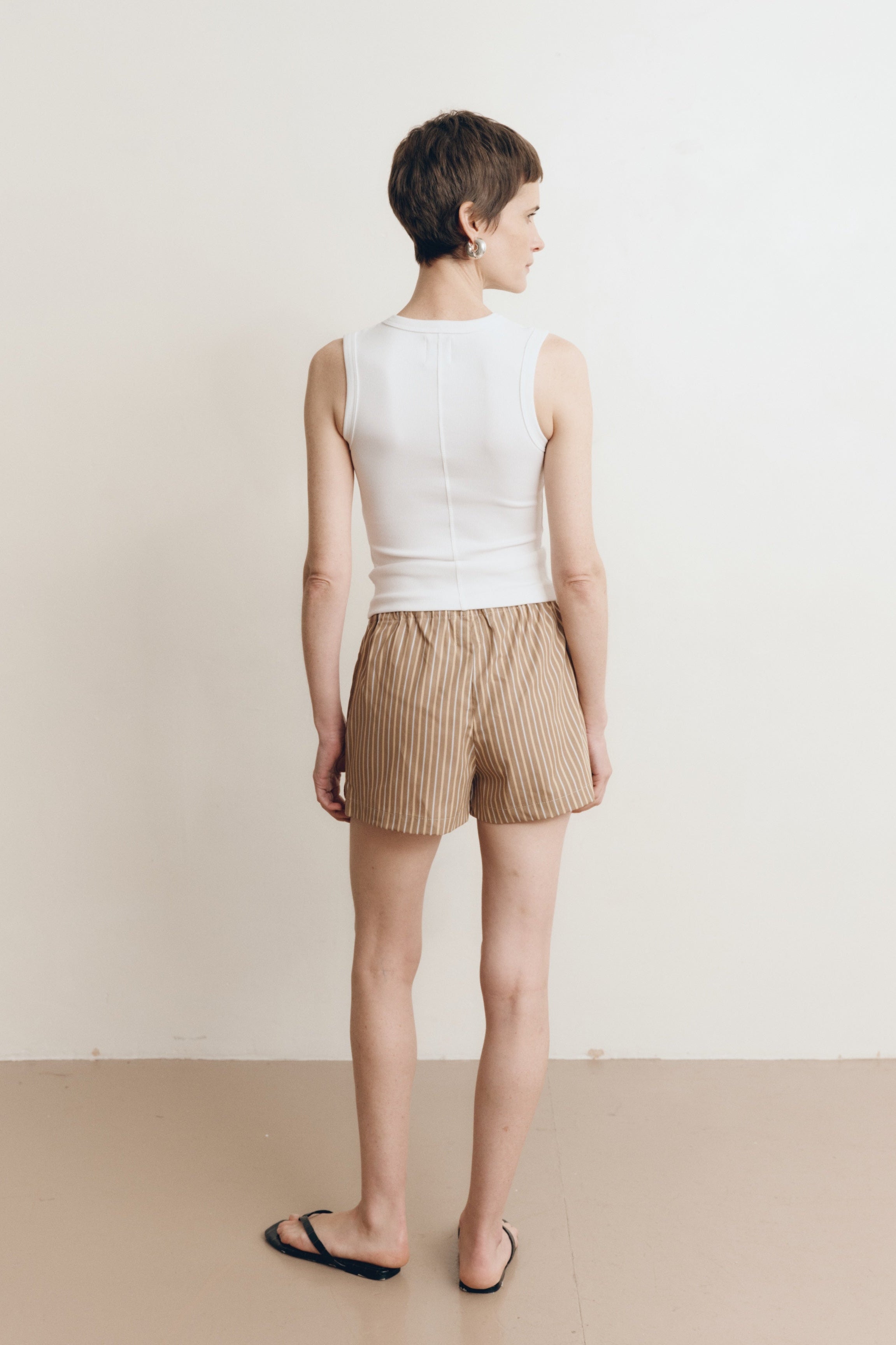 Posed female model wearing brown 100% cotton sustainable drawstring relaxed shorts paired with a high neck tank top in white and black sandals- VIR VITA
#color_canyon stripe