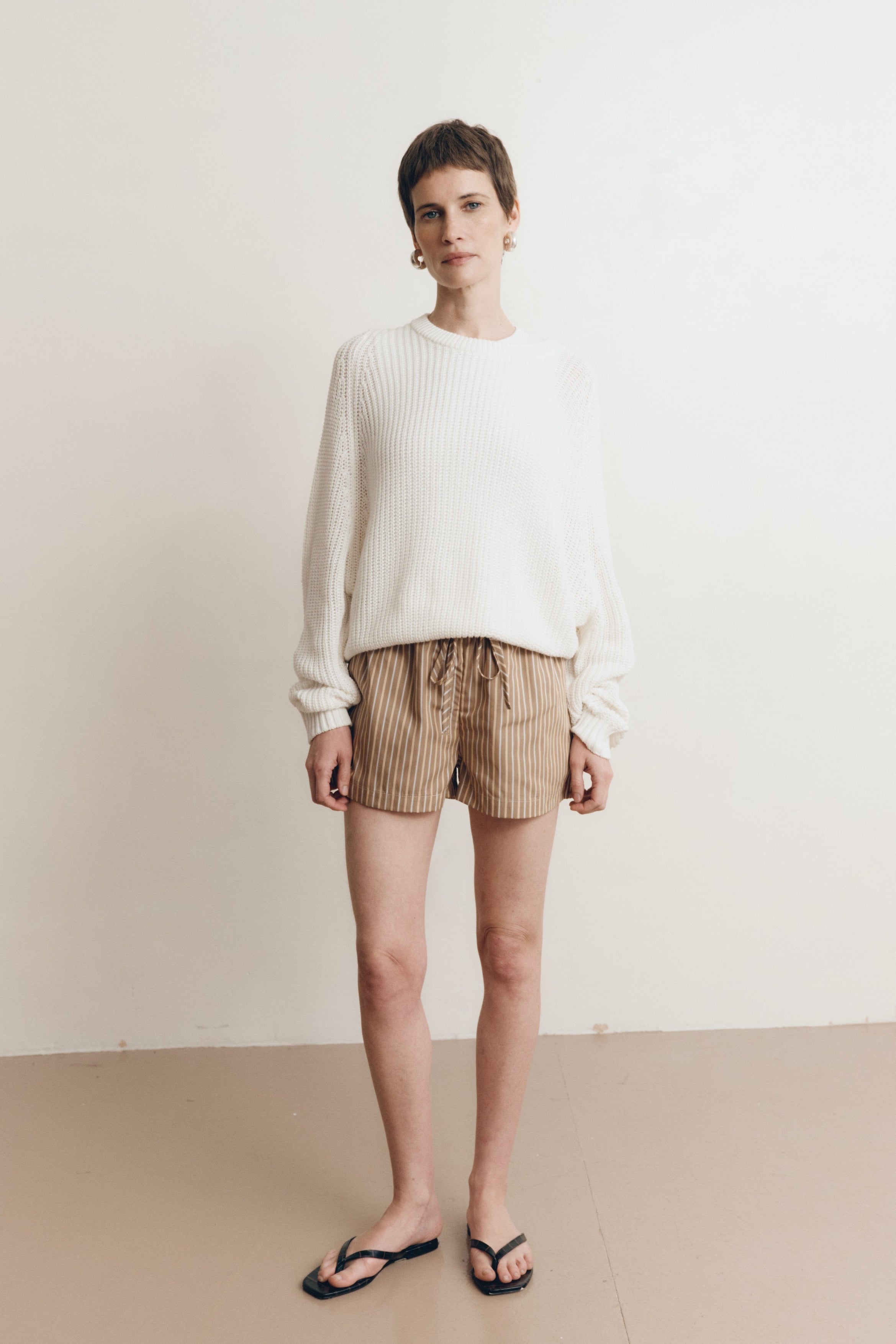 Posed female model wearing a 100% cotton knit sweater with a flattering and subtly cropped silhouette in white paired with striped brown drawstring shorts, and black sandals - VIR VITA
#color_bone