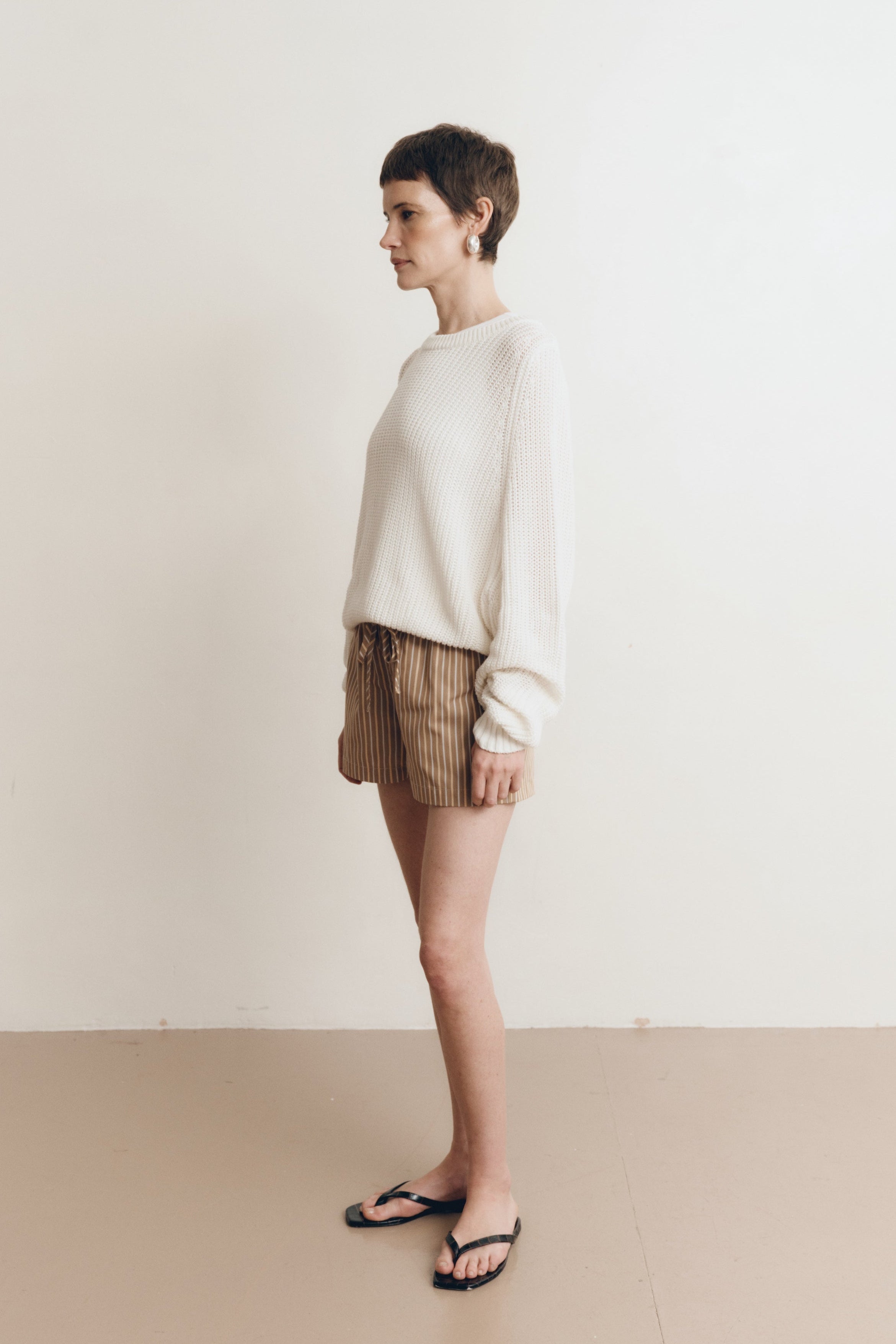 Posed female model wearing a 100% cotton knit sweater with a flattering and subtly cropped silhouette in white paired with striped brown drawstring shorts, and black sandals - VIR VITA
#color_bone