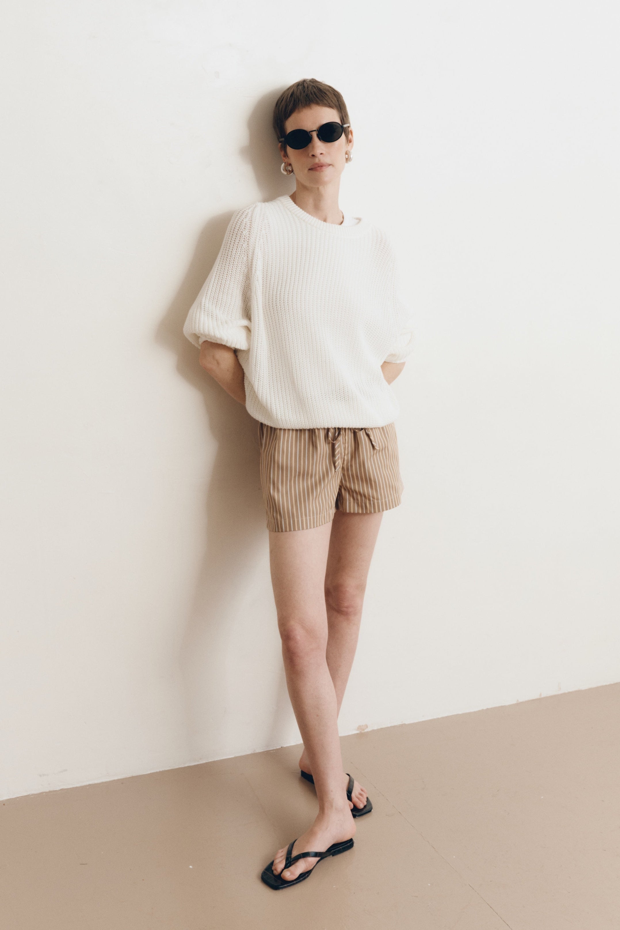 Posed female model wearing a 100% cotton knit sweater with a flattering and subtly cropped silhouette in white paired with striped brown drawstring shorts, black sandals, and black sunglasses - VIR VITA
#color_bone