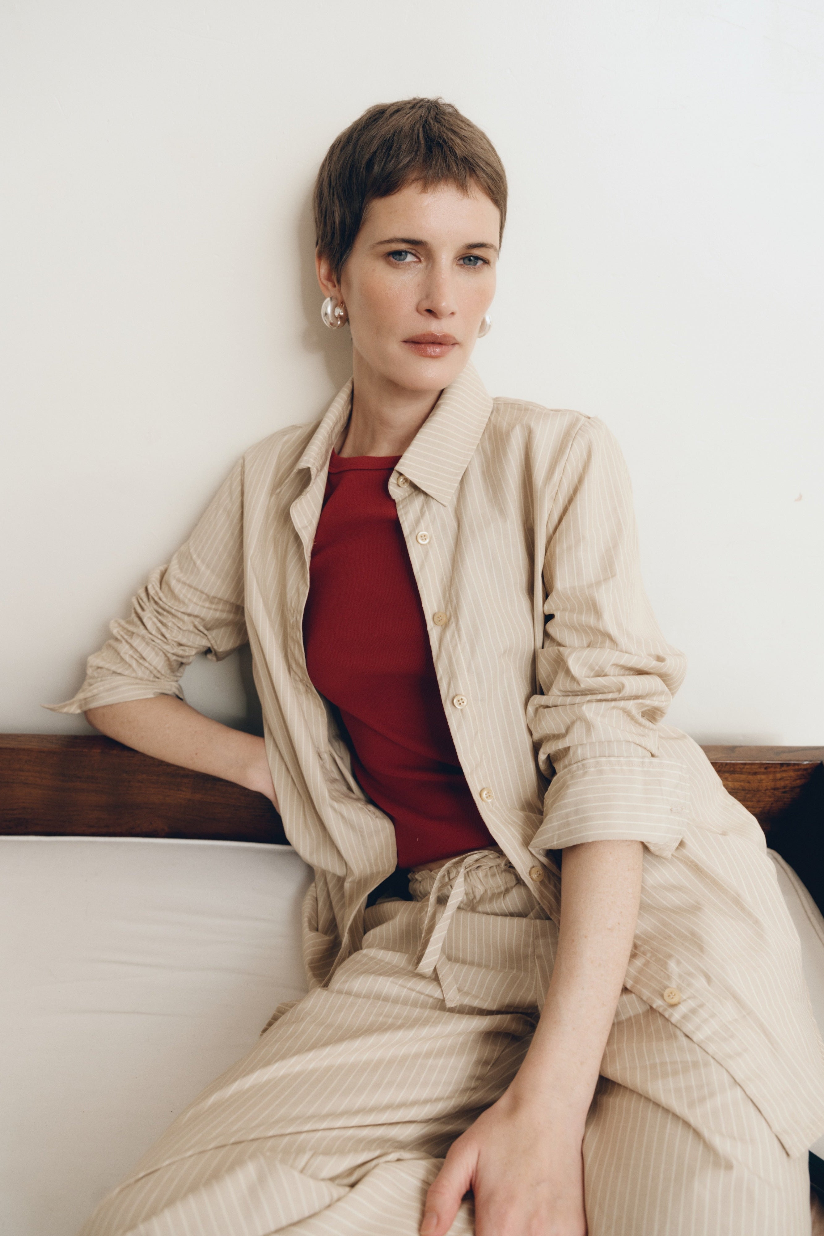 Posed female model sitting on a bench wearing a sustainable 100% cotton striped button up collared shirt in beige paired with matching striped drawstring pants and a high neck tank top in dark red - VIR VITA
#color_dune stripe