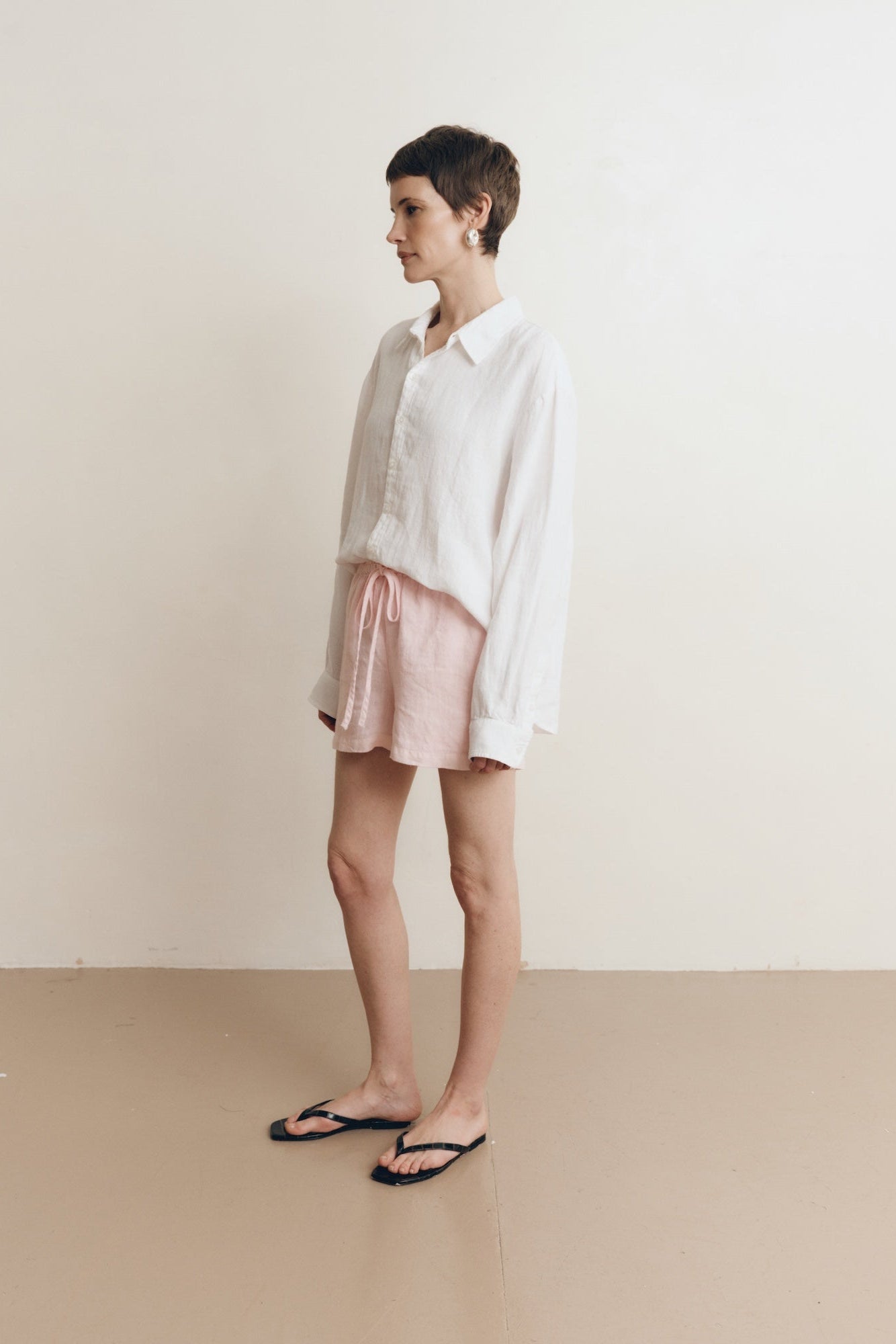 Posed female model wearing sustainable 100% linen drawstring shorts in light pink paired with a linen button up oversized shirt in white, and black sandals - VIR VITA
#color_blossom