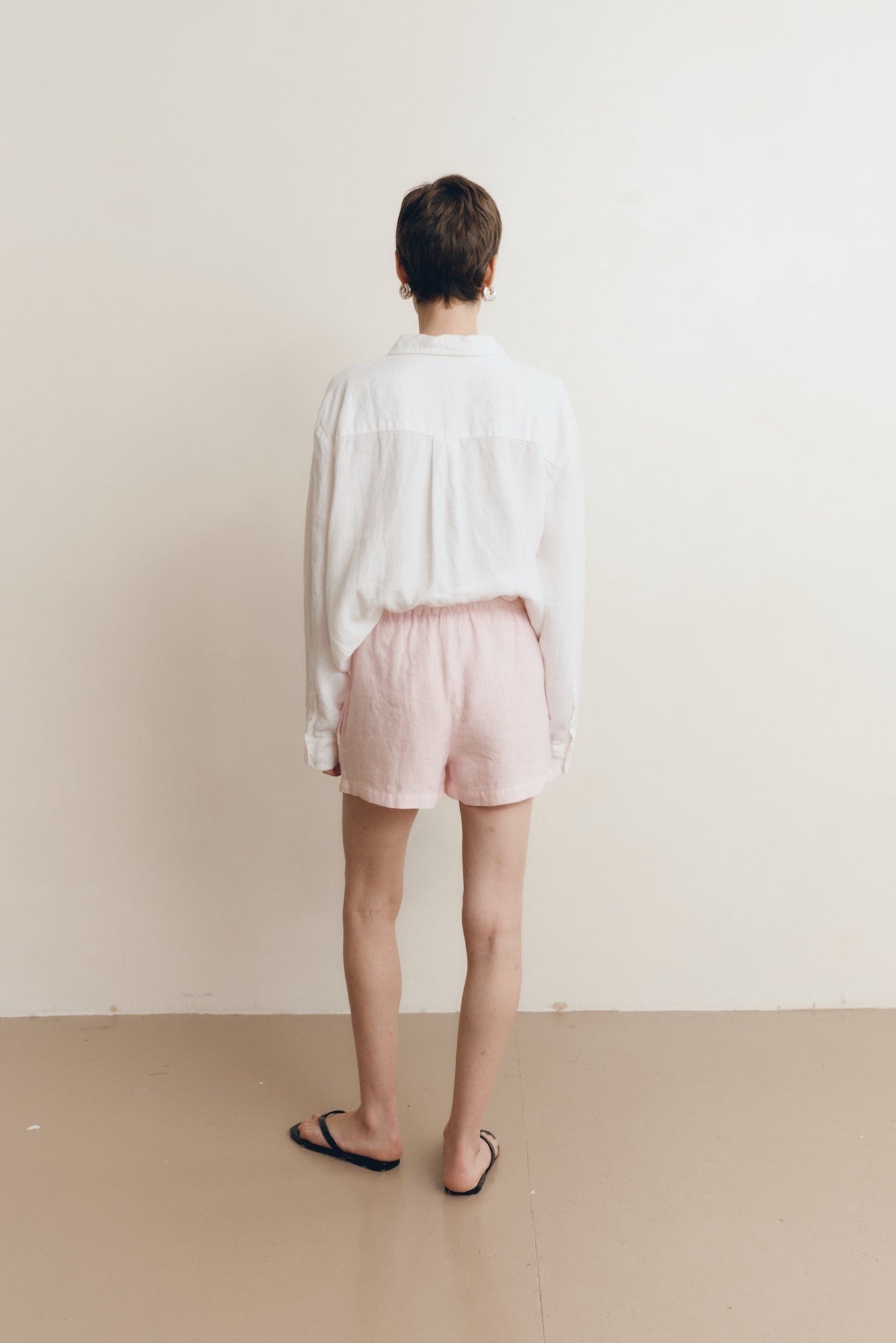 Posed female model wearing sustainable 100% linen drawstring shorts in light pink paired with a linen button up oversized shirt in white, and black sandals - VIR VITA
#color_blossom