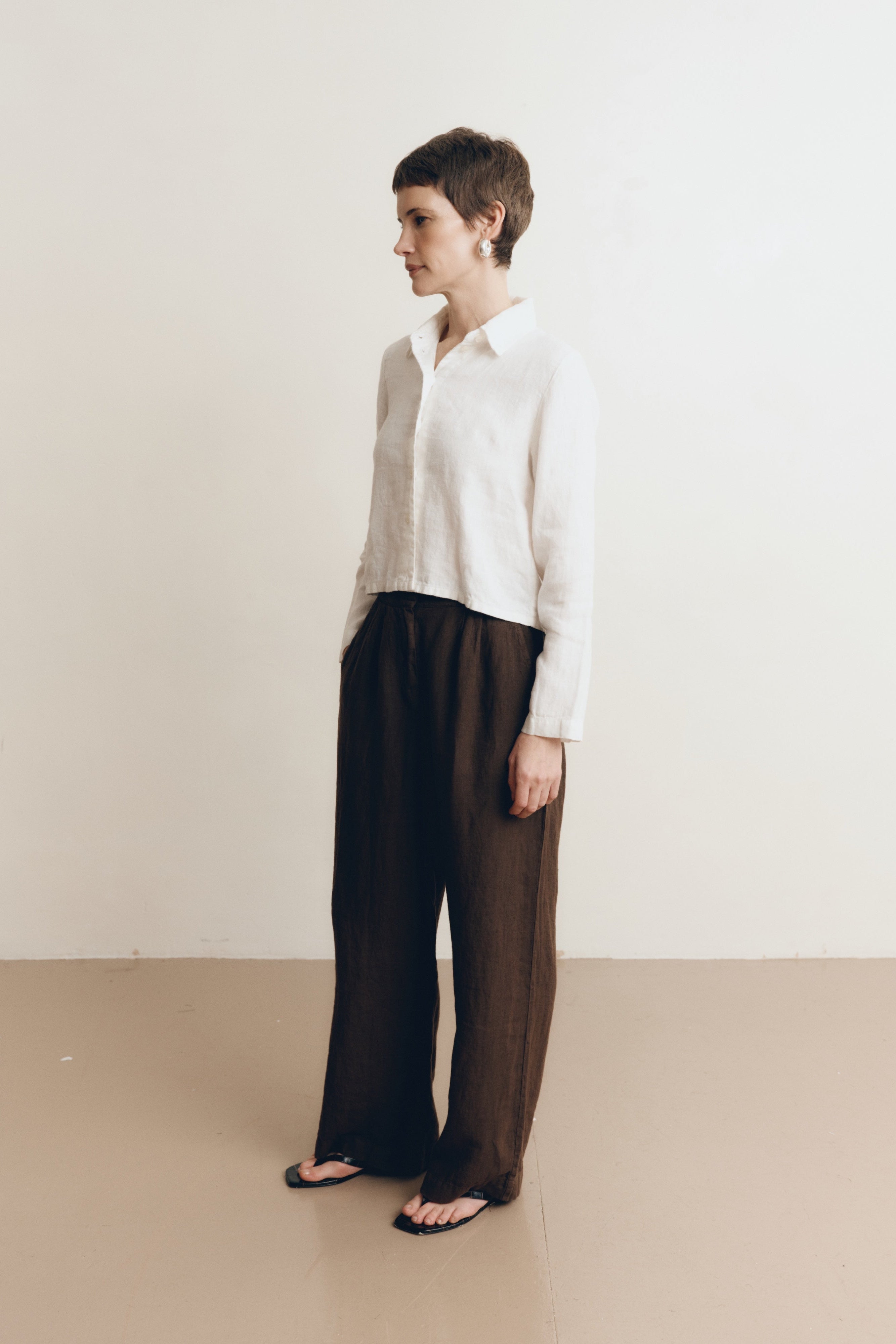 Posed female model wearing a sustainable 100% linen classic button up collared shirt in white paired with dark brown linen trousers and black sandals- VIR VITA
#color_white