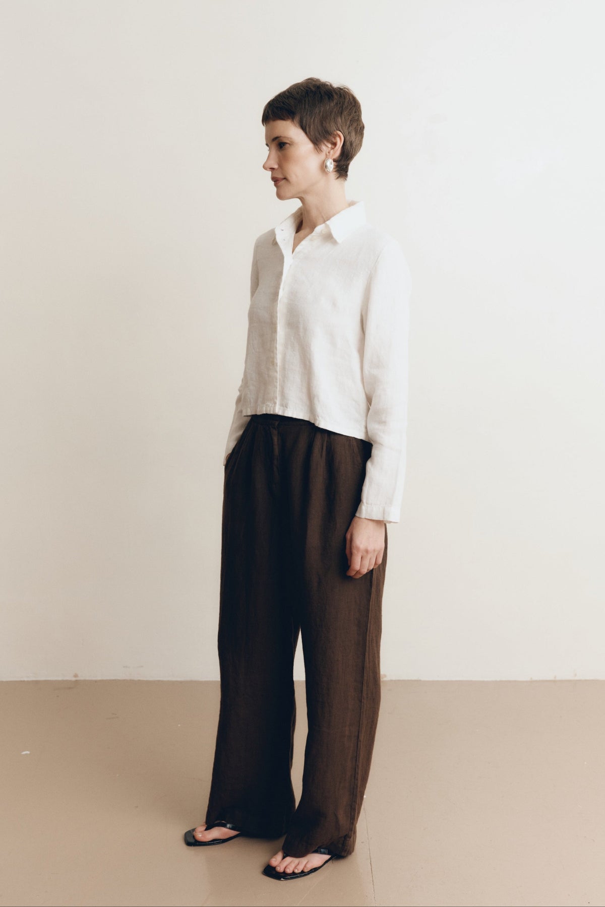 Posed female model wearing a sustainable 100% linen classic button up collared shirt in white paired with dark brown linen trousers and black sandals- VIR VITA
#color_white
