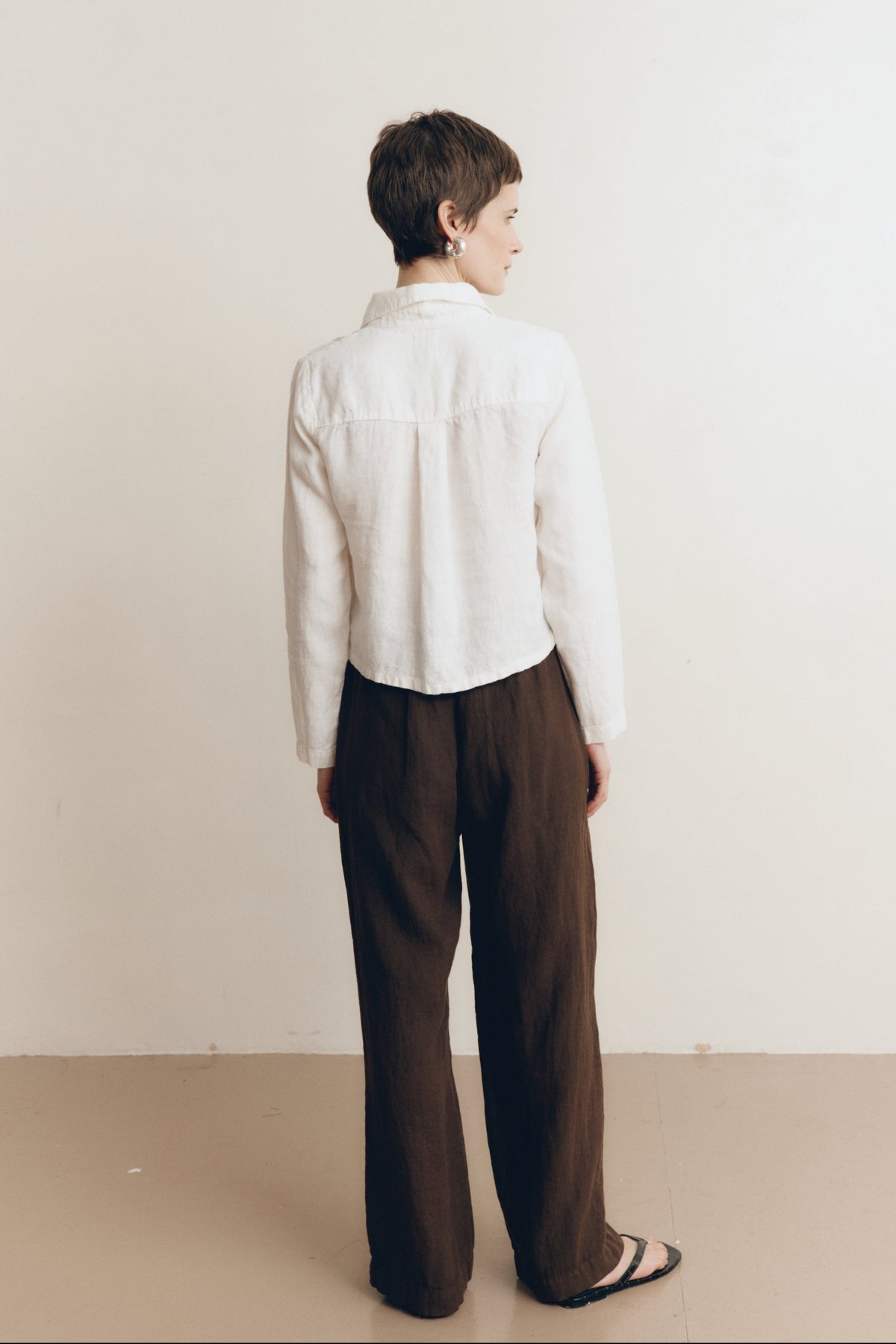 Posed female model wearing a sustainable 100% linen classic button up collared shirt in white paired with dark brown linen trousers and black sandals- VIR VITA
#color_white