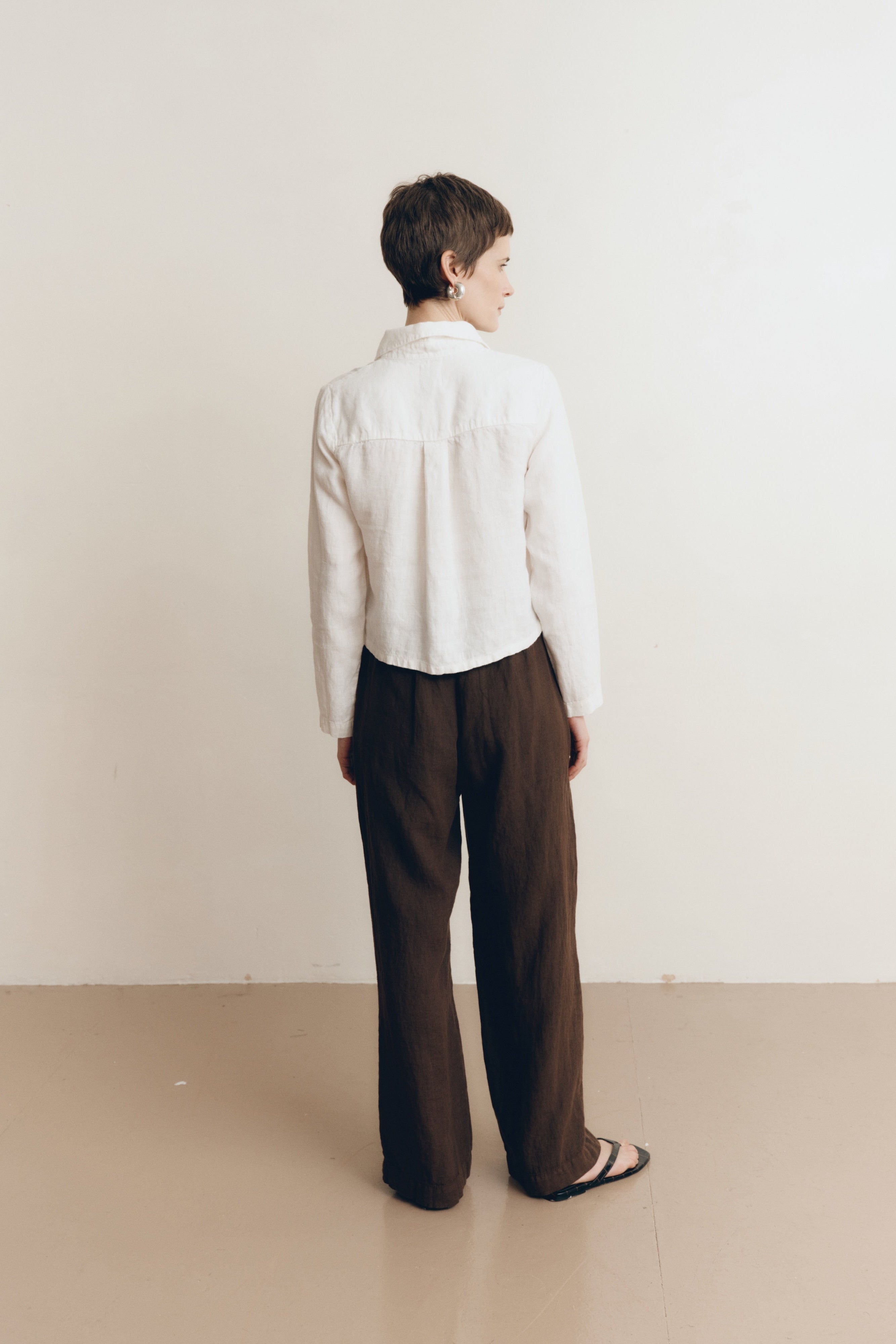 Posed female model wearing a sustainable 100% linen classic button up collared shirt in white paired with dark brown linen trousers and black sandals- VIR VITA
#color_white
