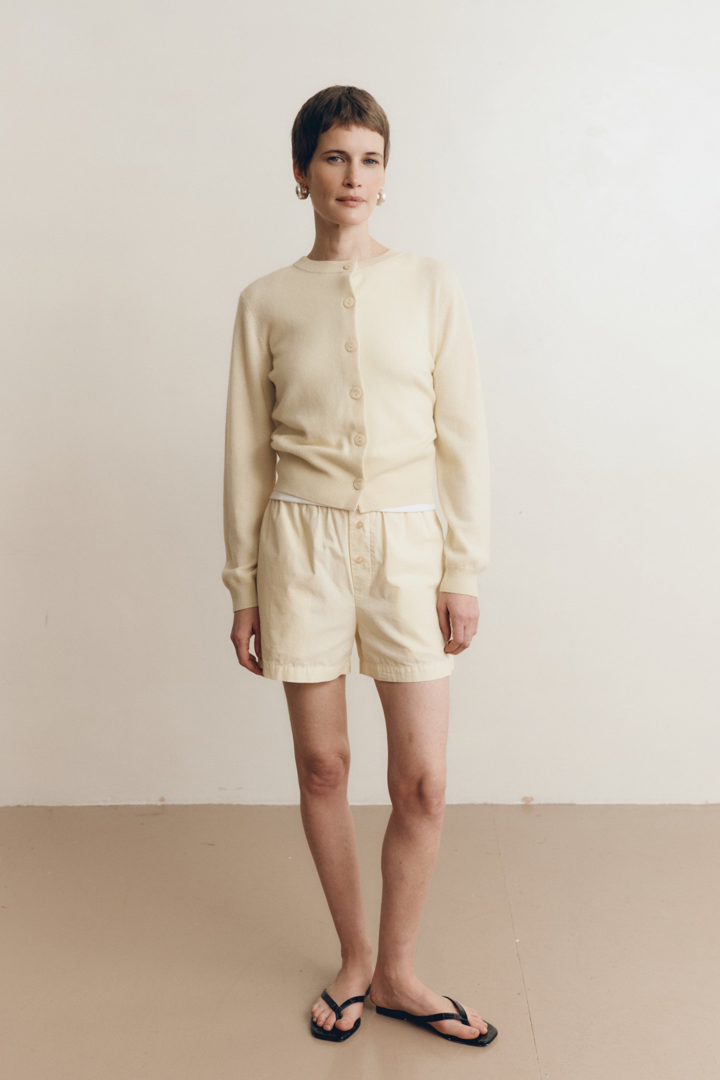 Posed female model wearing 100% cotton poplin relaxed shorts with a button closure in pale yellow paired with a pale yellow matching cashmere cardigan and black sandals - VIR VITA
#color_butter