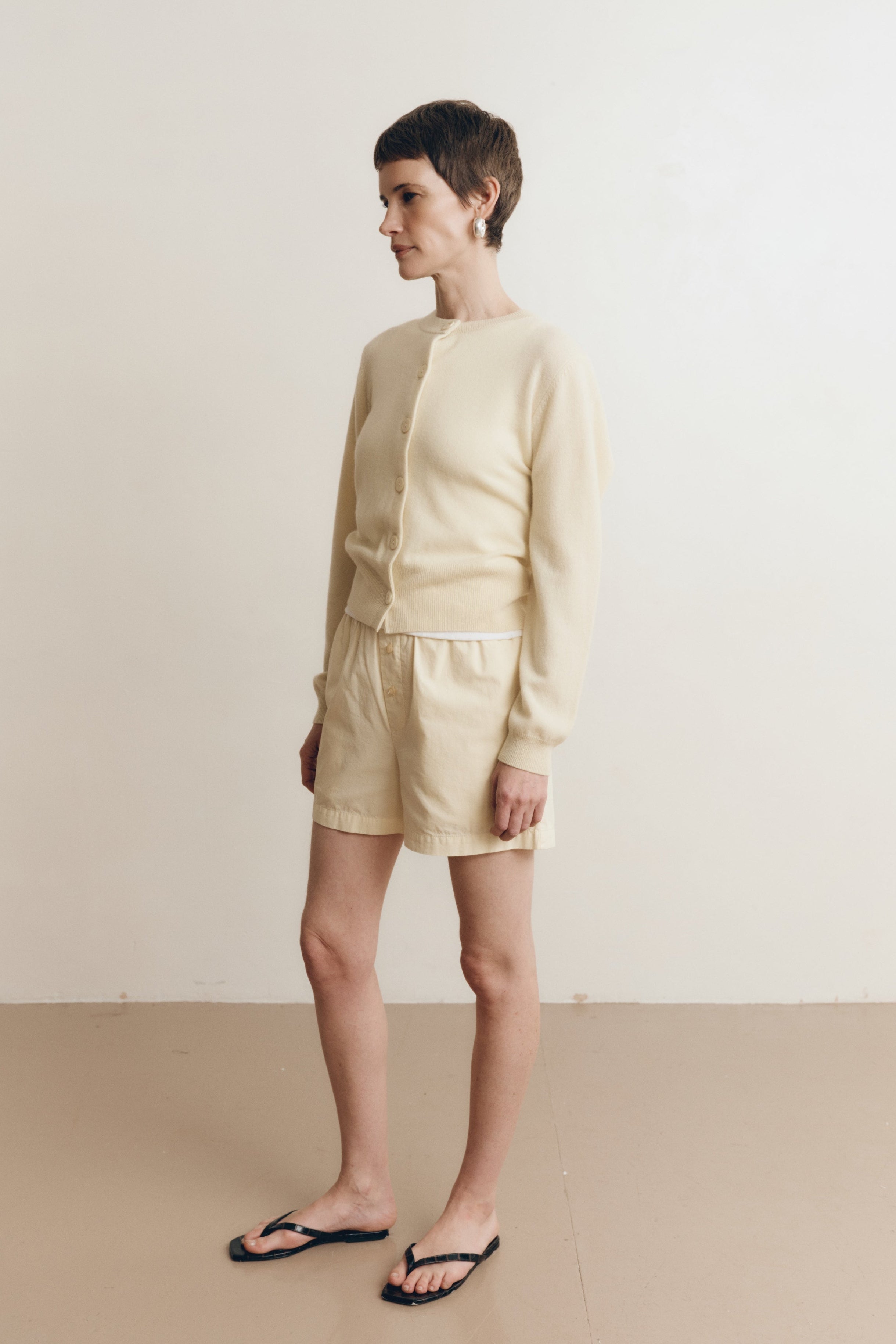 Posed female model wearing 100% cotton poplin relaxed shorts with a button closure in pale yellow paired with a pale yellow matching cashmere cardigan and black sandals - VIR VITA
#color_butter