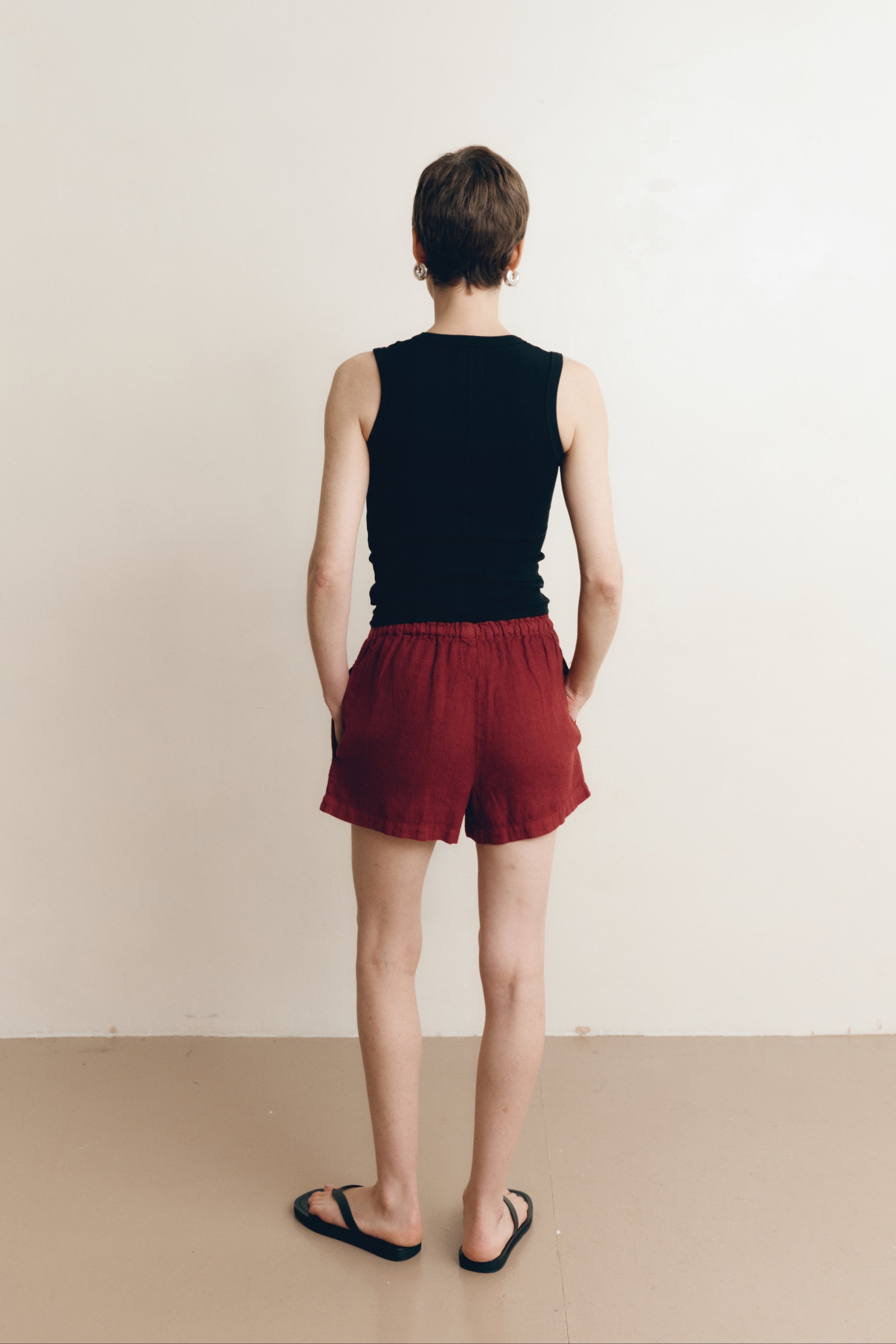 Posed female model wearing sustainable 100% linen drawstring shorts in dark red paired with a black v-neck tank top, and black sandals - VIR VITA
#color_sun dried tomato