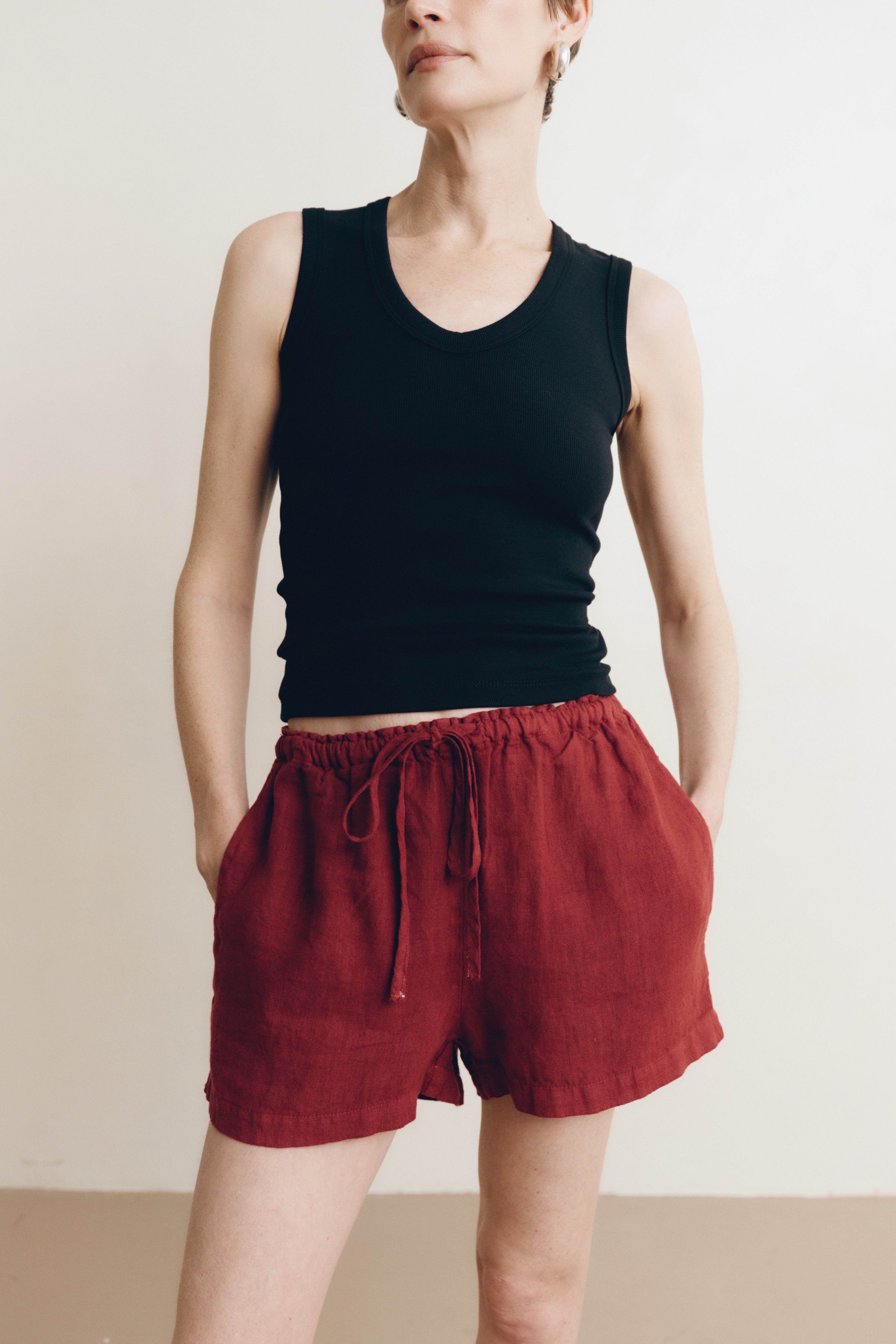 Posed female model wearing sustainable 100% linen drawstring shorts in dark red paired with a black v-neck tank top - VIR VITA
#color_sun dried tomato