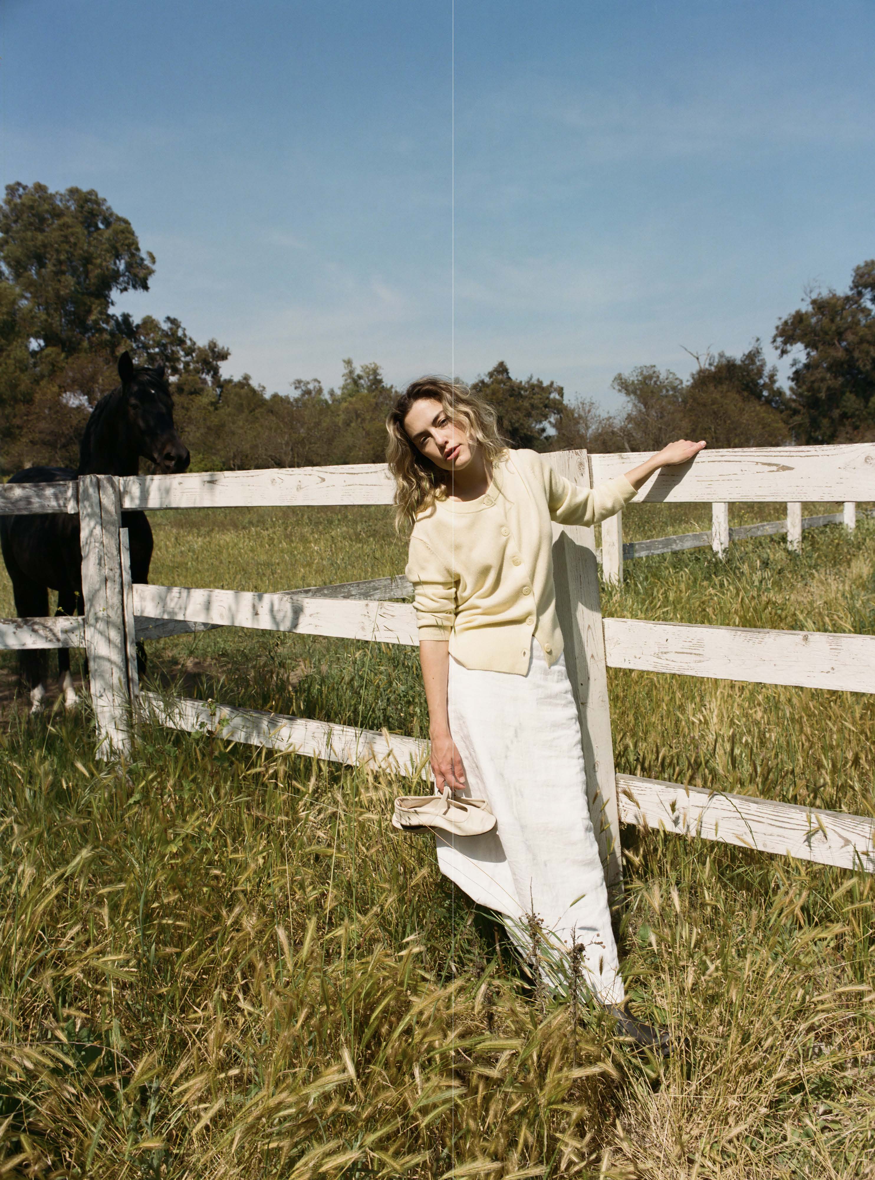 Female model leaning against a fence wearing a pale yellow ultra-soft luxurious cashmere button up cardigan paired with a white linen maxi skirt, and white flats - VIR VITA
#color_butter