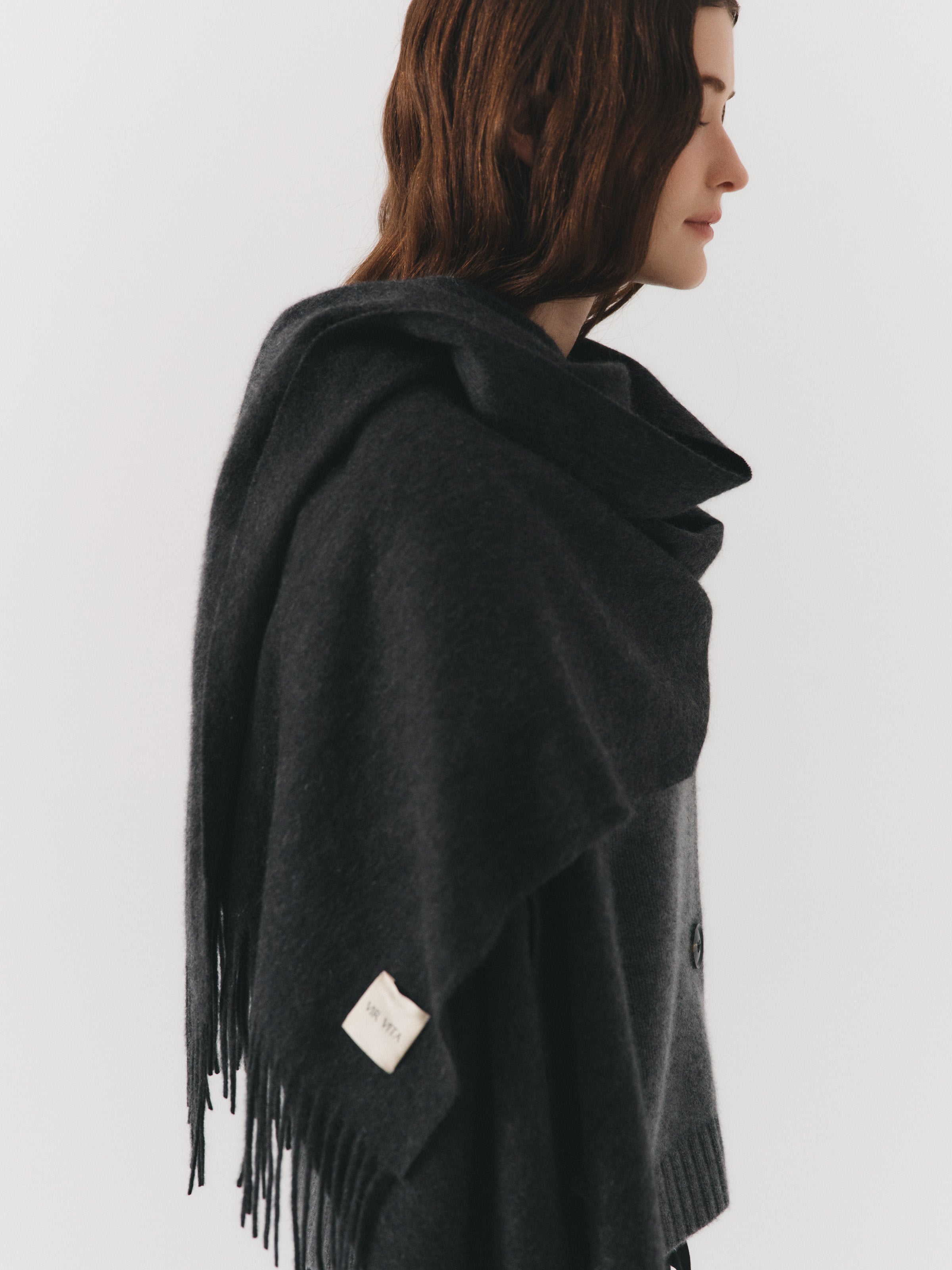 Female model posing wearing a dark grey brushed cashmere scarf with a fringed edge at both ends. Cut long, oversized enough to wrap or drape over the shoulder. It is paired with a cashmere cardigan in stone grey. - VIR VITA
#color_anthracite