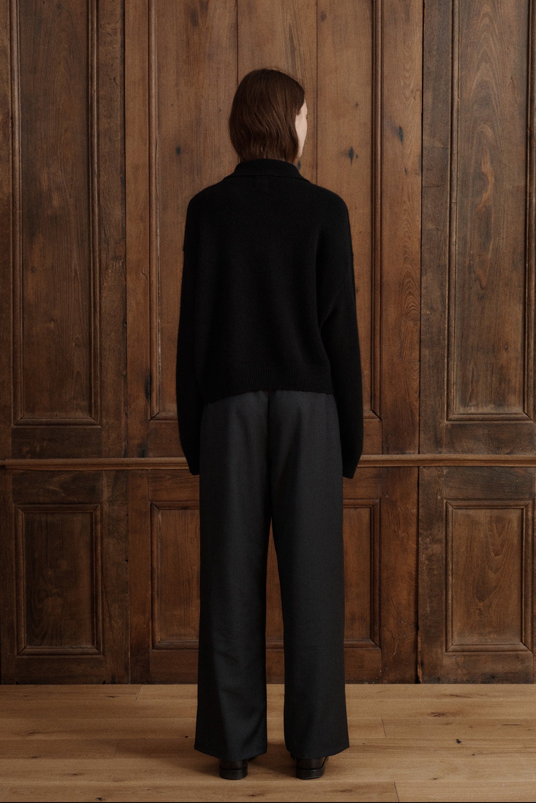 Female model posing wearing a black polo sweater crafted from soft cashmere. Designed with a structured collar, shoulder, and four-button front closure for a timeless look. Paired with dark grey wool elastic waist trousers and black loafers - VIR VITA 
#color_black