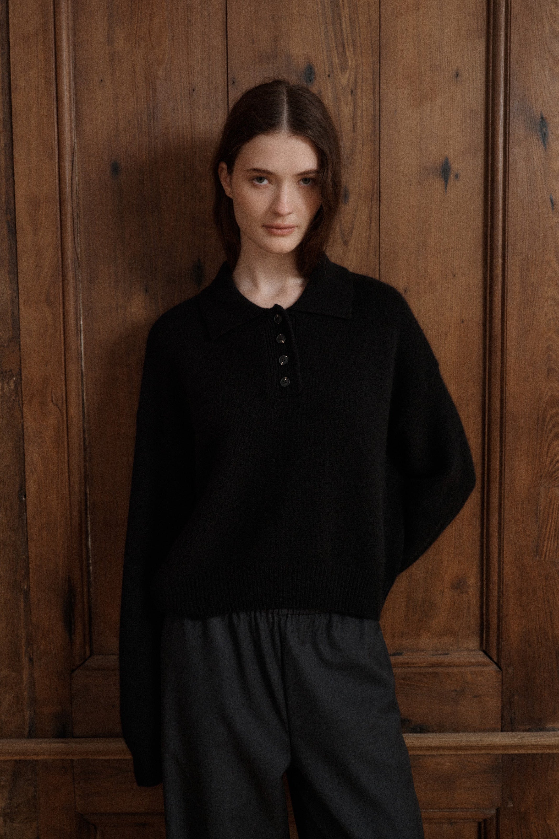 Female model posing wearing a black polo sweater crafted from soft cashmere. Designed with a structured collar, shoulder, and four-button front closure for a timeless look. Paired with dark grey wool elastic waist trousers - VIR VITA 
#color_black