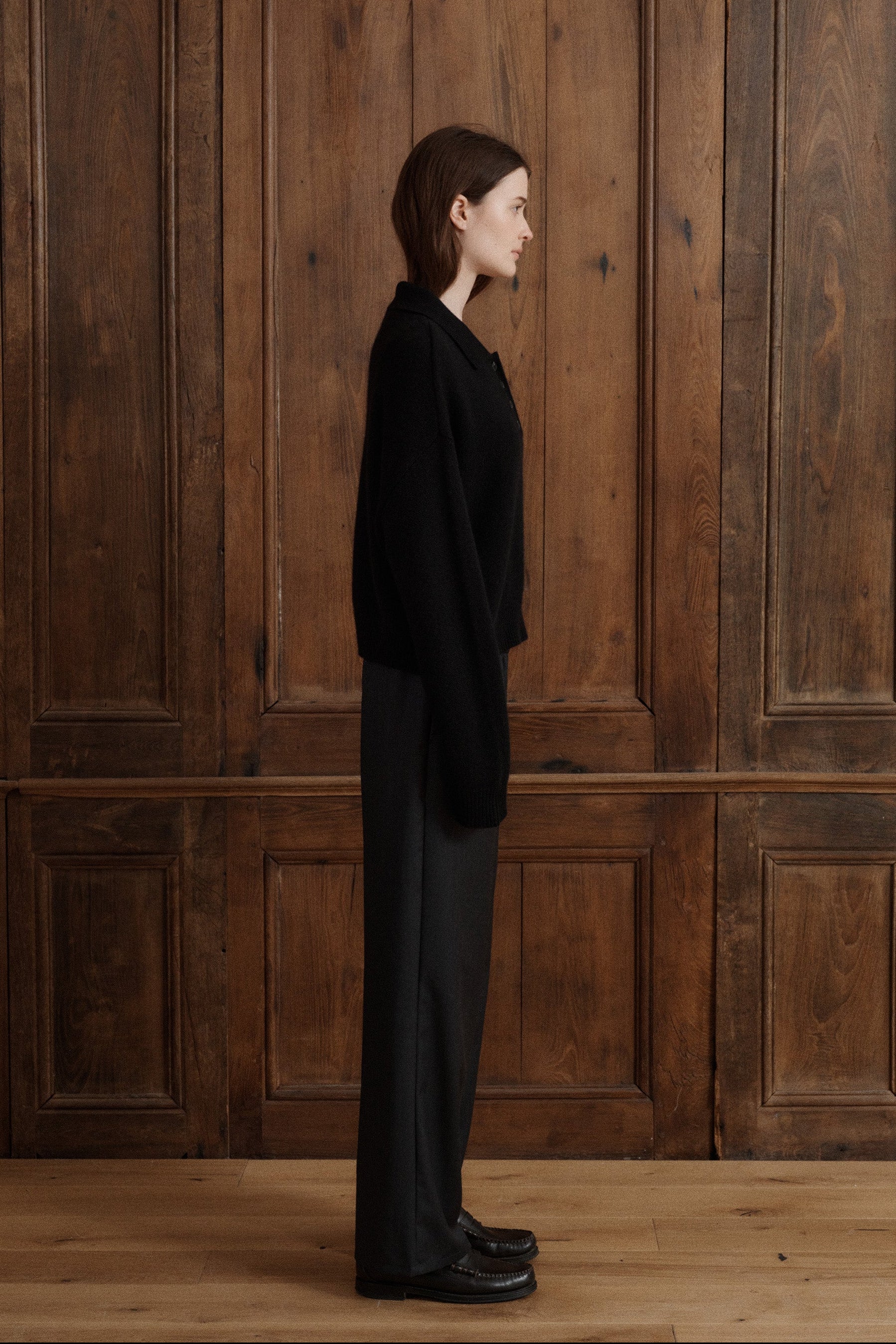 Female model posing wearing a black polo sweater crafted from soft cashmere. Designed with a structured collar, shoulder, and four-button front closure for a timeless look. Paired with dark grey wool elastic waist trousers and black loafers - VIR VITA 
#color_black