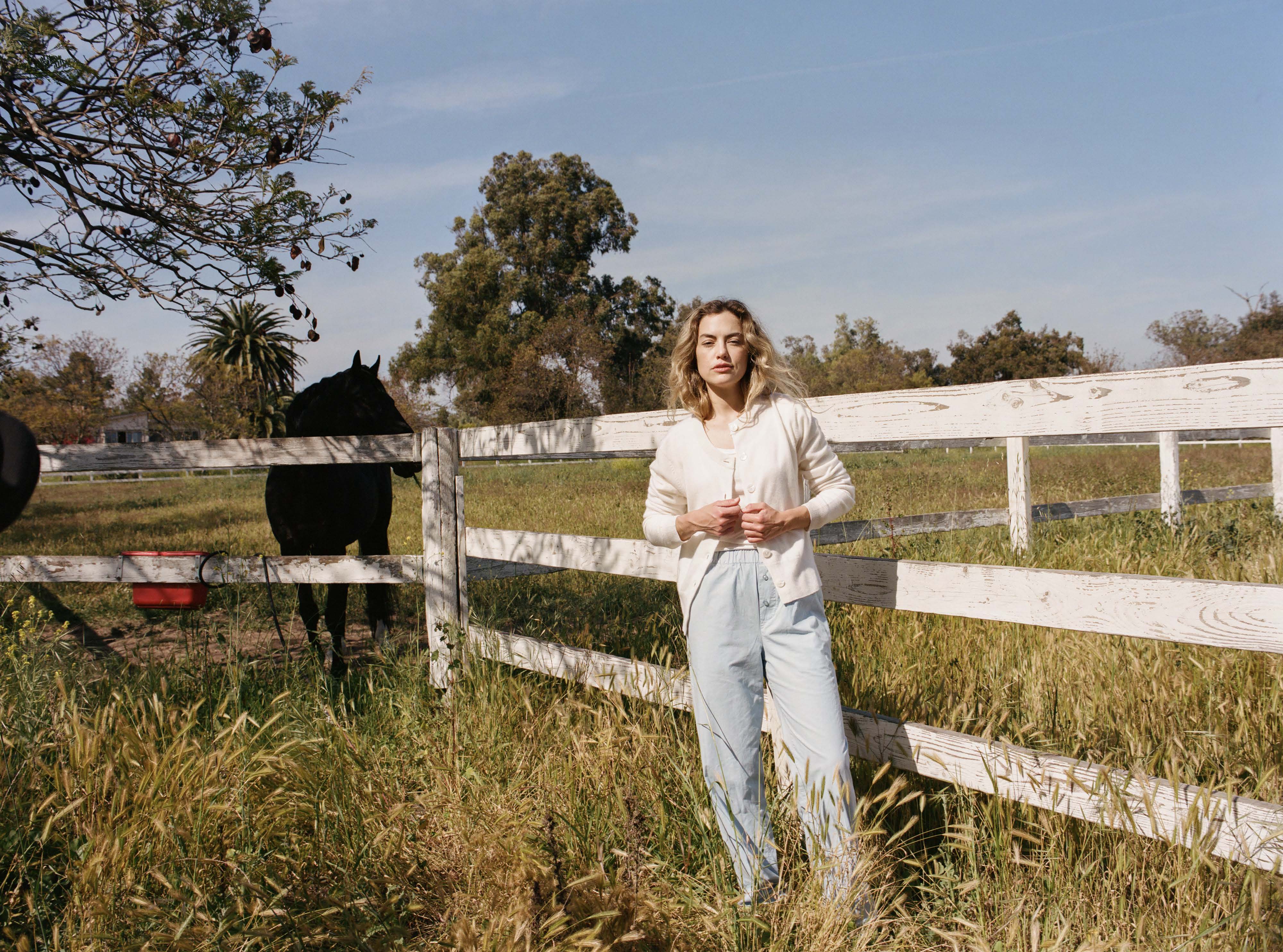 Female model standing next to a fence wearing a white ultra-soft luxurious cashmere button up cardigan paired with light blue cotton poplin pants - VIR VITA
#color_bone