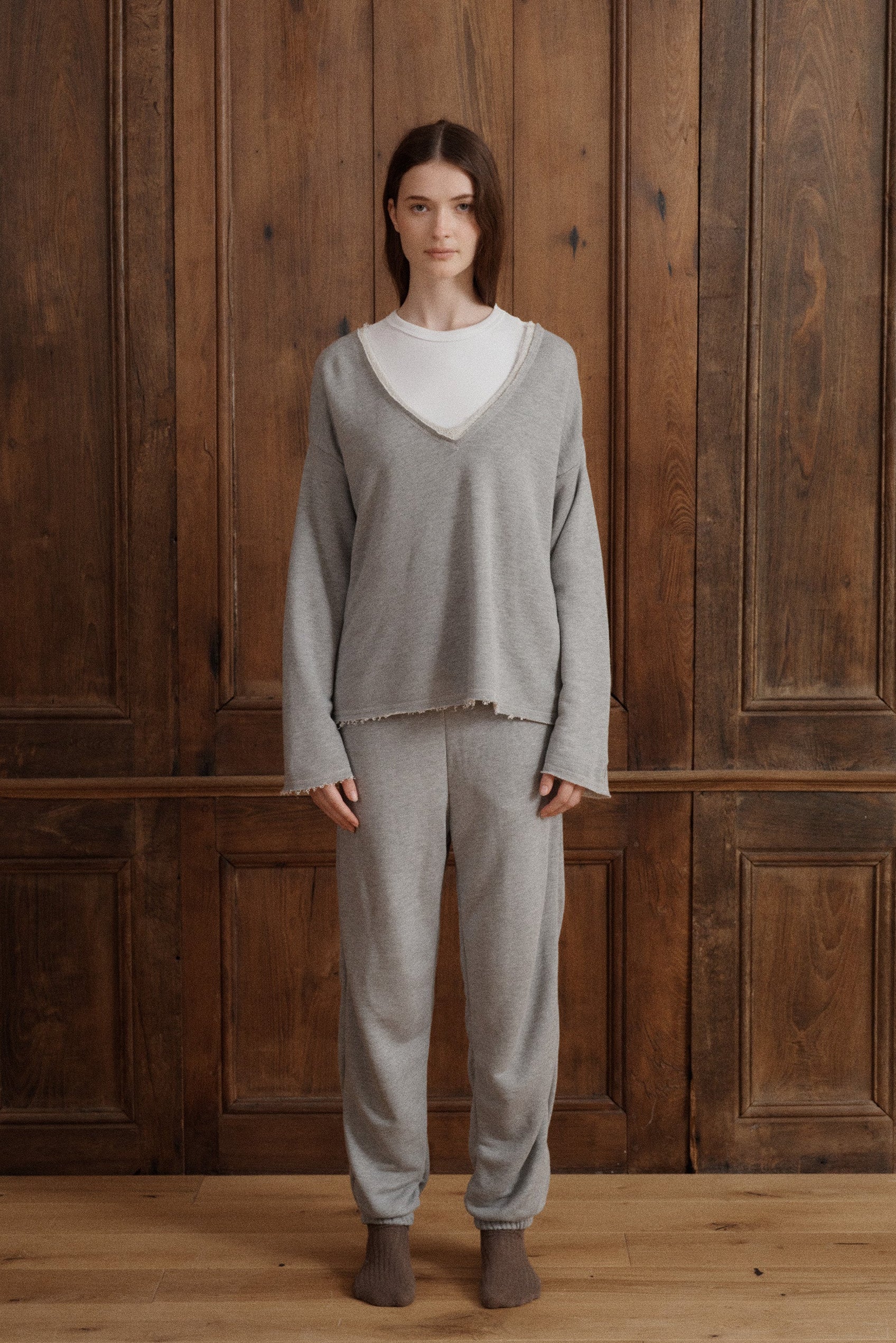 Female model wearing a matching heather grey loungewear set featuring a v-neck relaxed sweatshirt with a raw hem, relaxed joggers cinched at the waist and ankles and brown socks. - VIR VITA
#color_heather grey