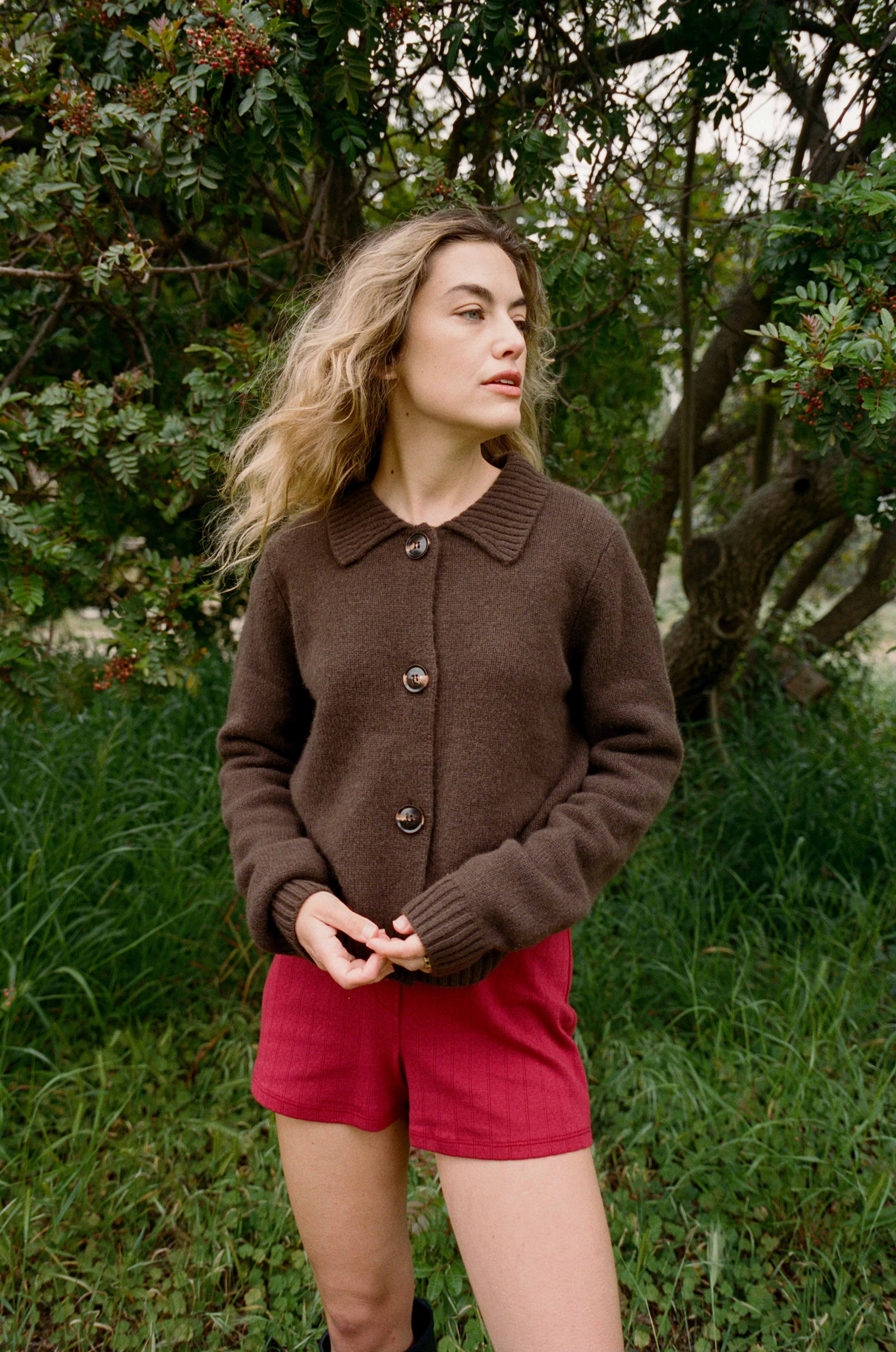 Female model standing in grass wearing a dark brown ultra-soft luxurious cashmere cardigan featuring a classic silhouette with a collar and large buttons paired with red pointelle shorts - VIR VITA
#color_espresso