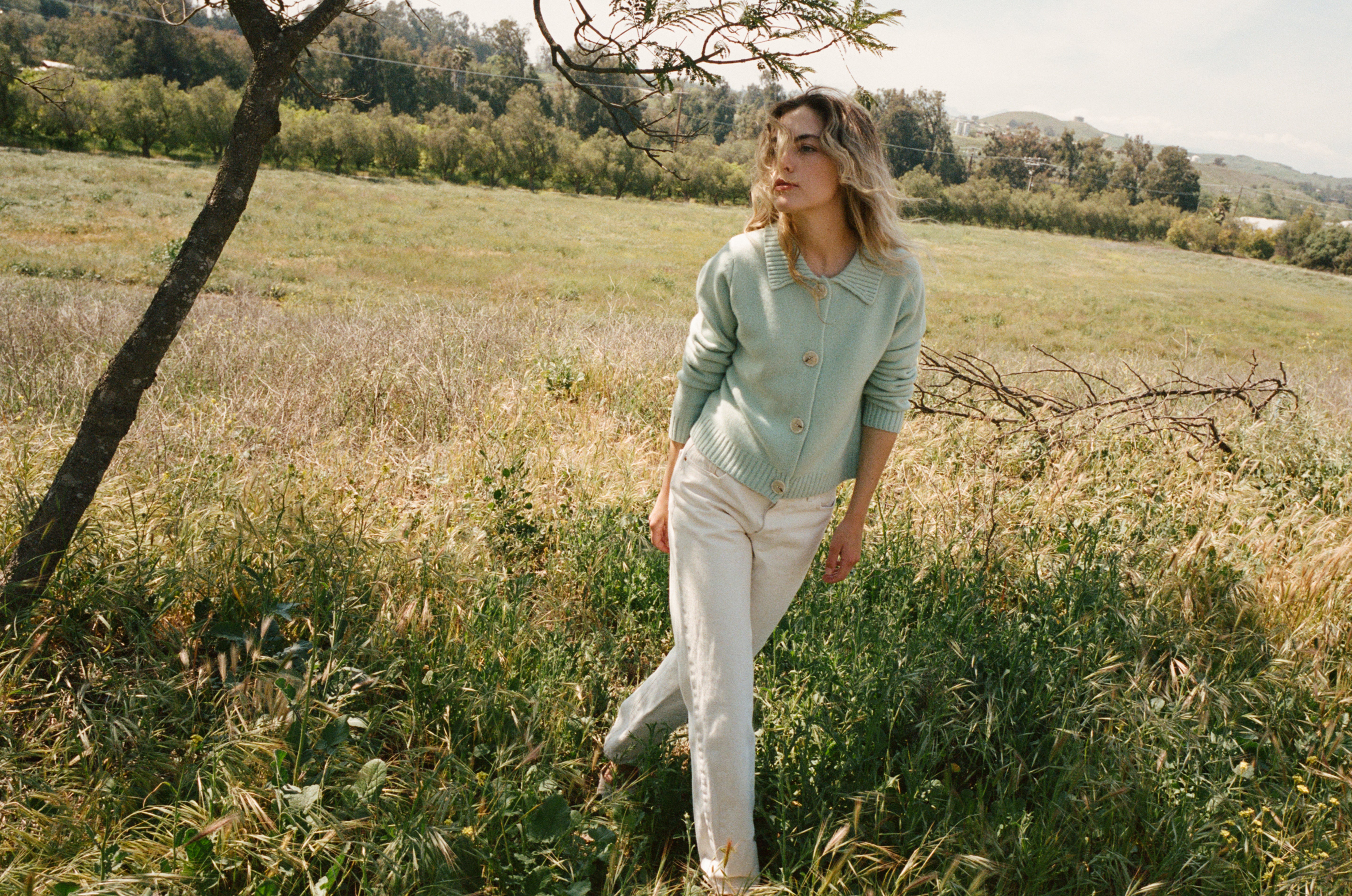 Female model standing in grass wearing a teal blue ultra-soft luxurious cashmere cardigan featuring a classic silhouette with a collar and large buttons paired with white jeans- VIR VITA
#color_sea glass