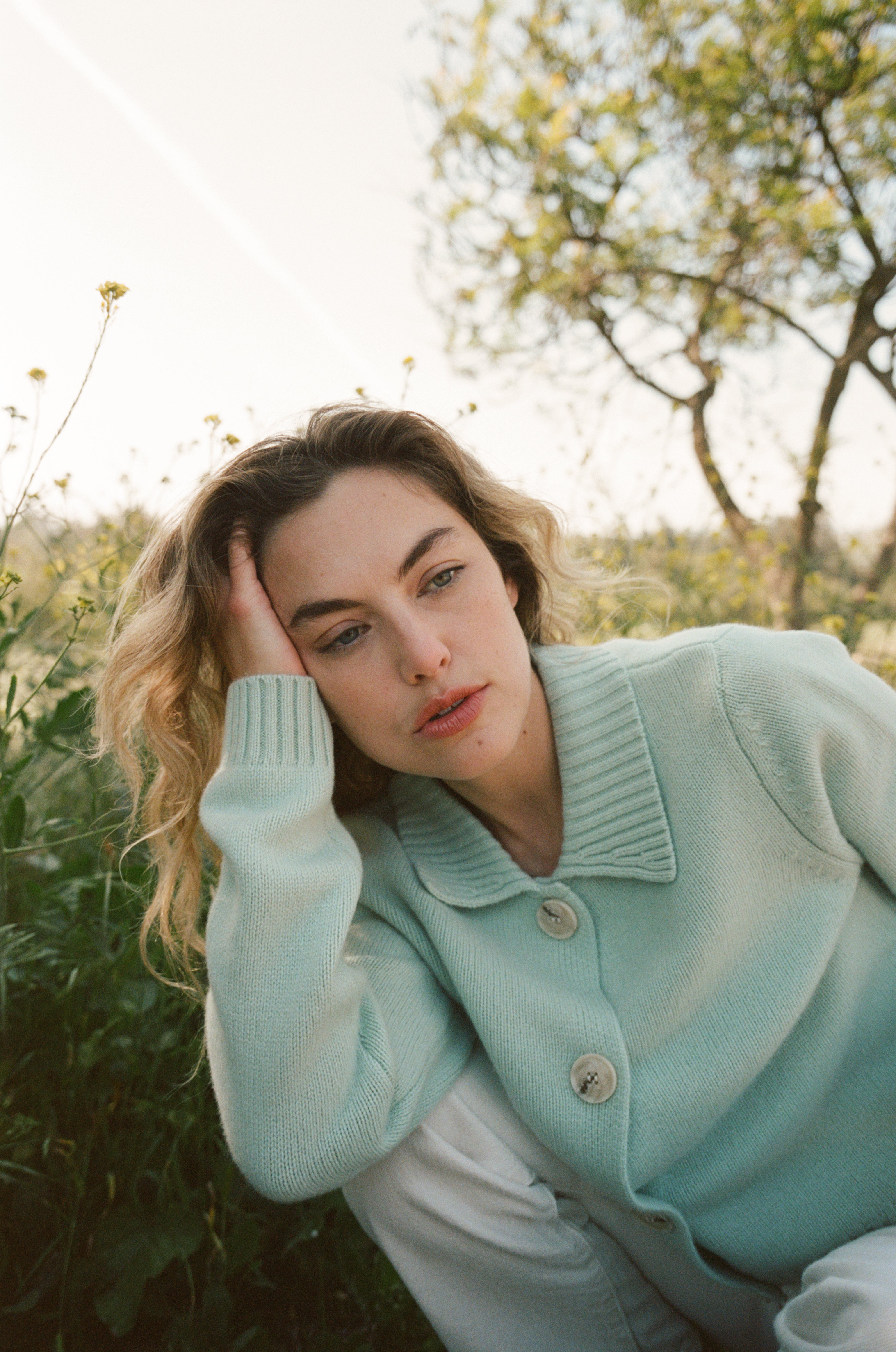 Female model sitting in grass wearing a teal blue ultra-soft luxurious cashmere cardigan featuring a classic silhouette with a collar and large buttons paired with white jeans- VIR VITA
#color_sea glass